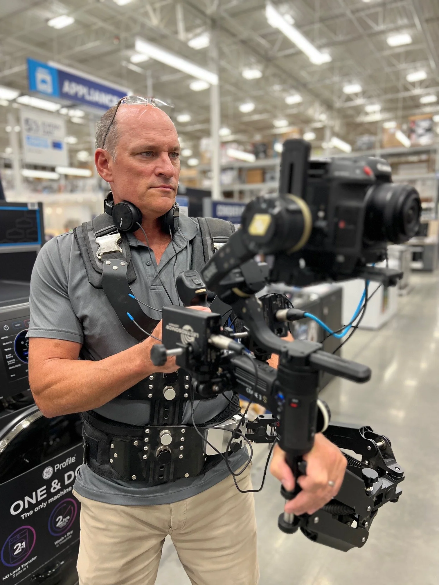A man operating a professional camera mounted on a stabilizer rig inside a retail store, with shelves and a sign reading 'APPAREL' in the background.