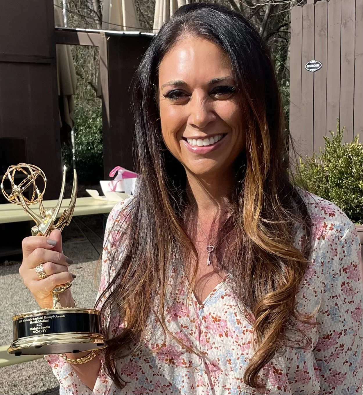 A woman with long brown hair smiling and holding an Emmy award outside, wearing a pink and white floral blouse and a necklace.