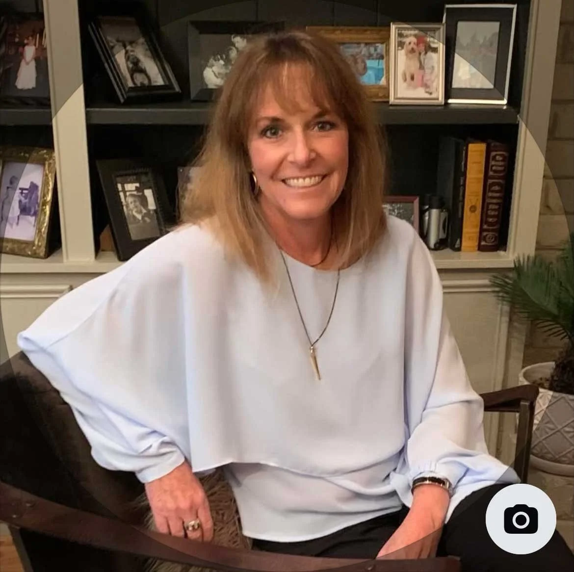 A smiling middle-aged woman with shoulder-length light brown hair sitting in a room with a bookshelf behind her, decorated with family photos, books, and framed pictures.