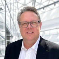 A man with glasses and gray hair, wearing a dark suit and white shirt, standing inside a modern glass building.