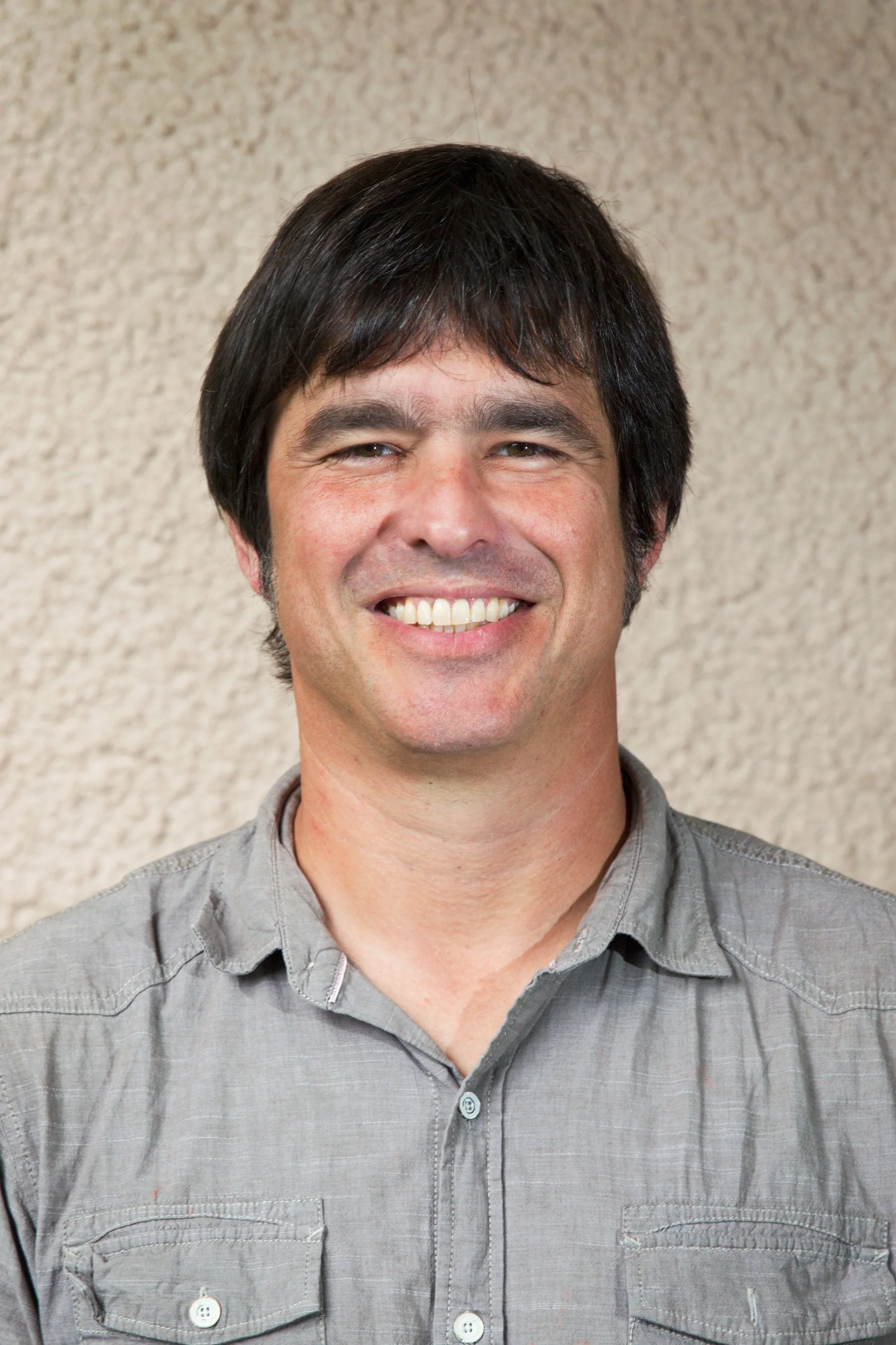 A smiling man with dark hair wearing a gray button-up shirt, standing in front of a textured beige wall.