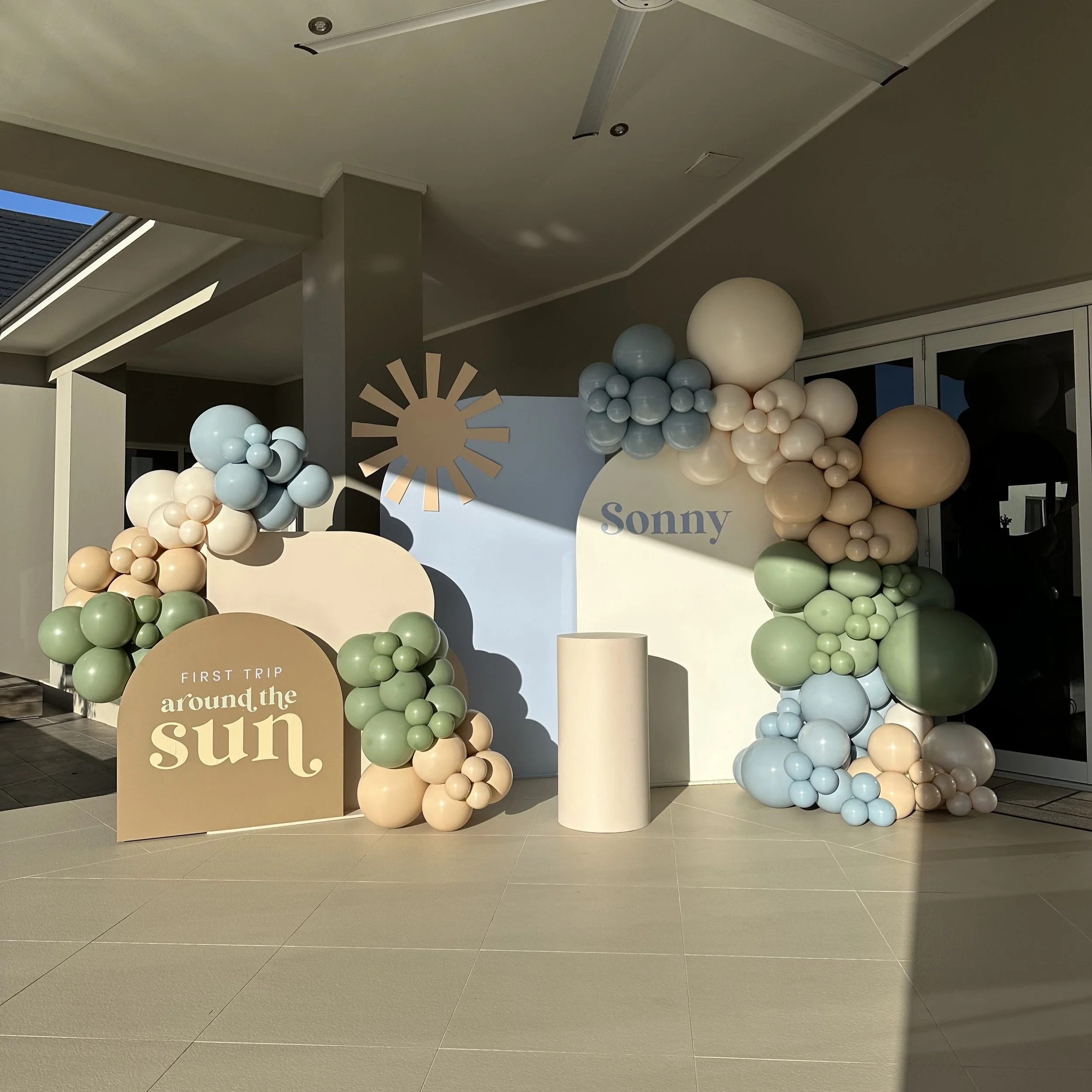 Decorative setup with pastel-colored balloons arranged around a backdrop for a first trip celebration. The backdrop has the name 'Sonny' and a sign reads 'First trip around the sun.' The scene is outdoors, with sunlight casting shadows.