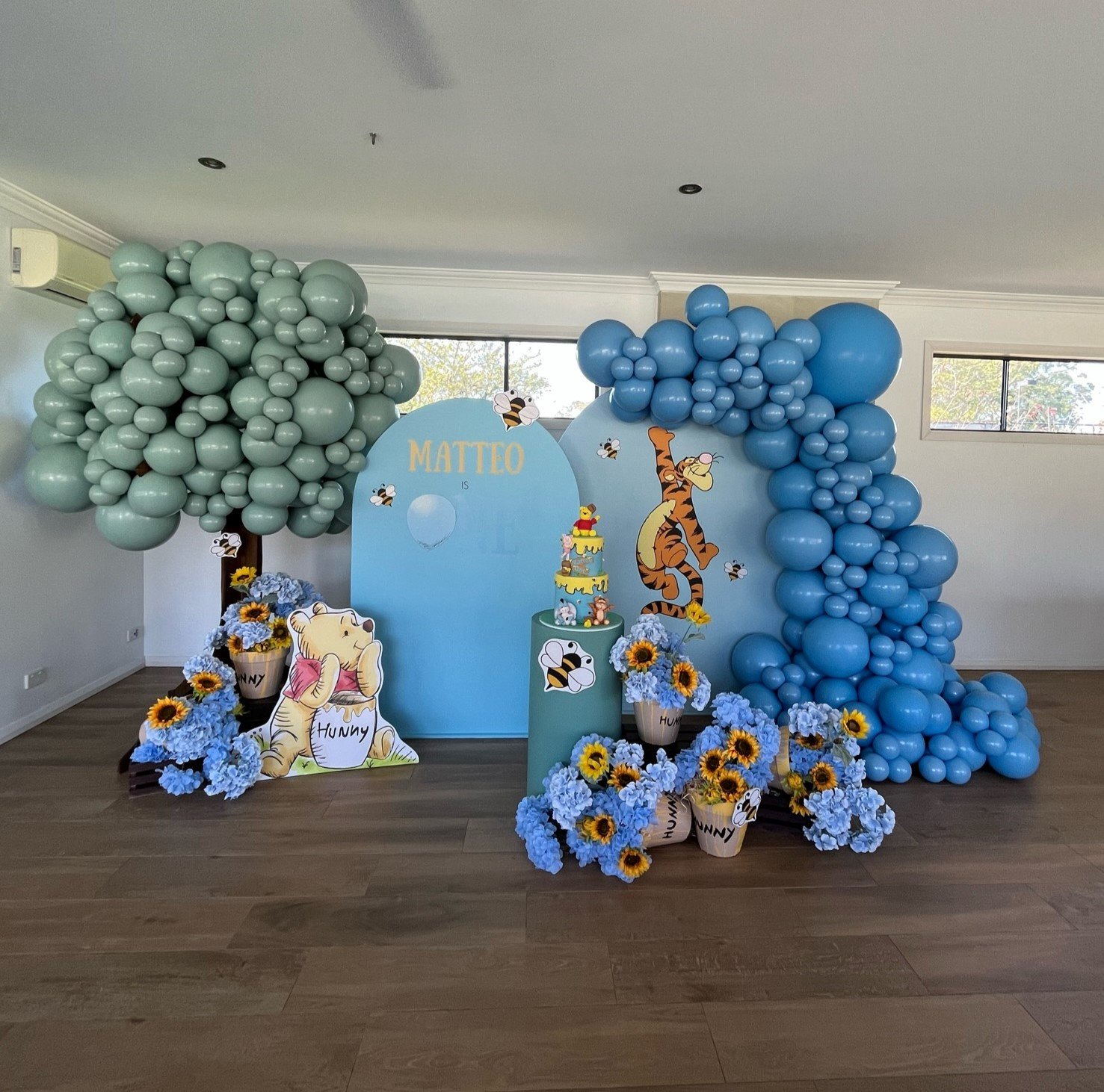 Celebration setup with balloon trees, a blue backdrop with Winnie the Pooh theme, and colorful flower arrangements for a child's birthday party.