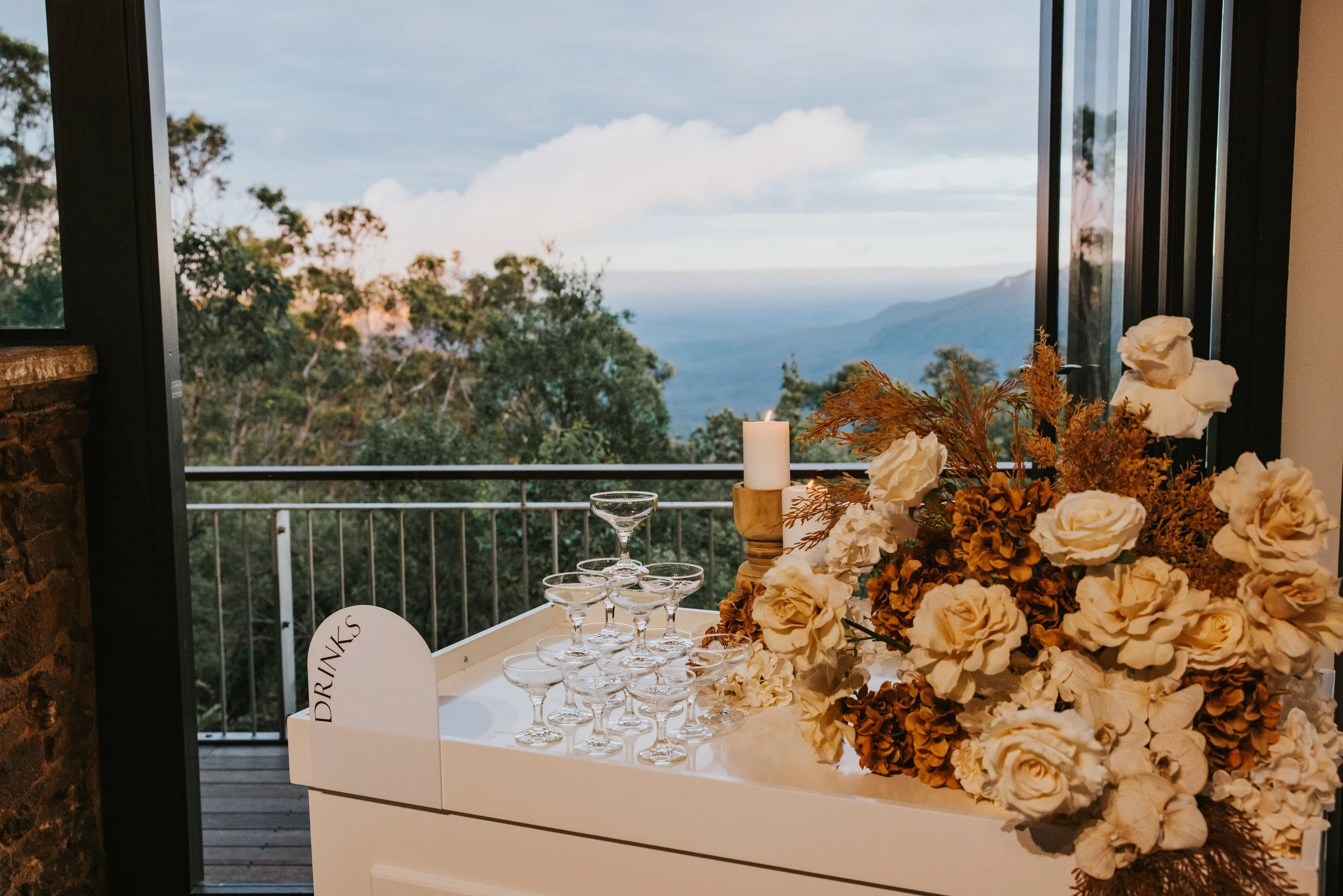 A tray with champagne glasses, a candle, and a large floral arrangement with white roses and dried foliage, set against a window overlooking a mountain landscape with trees and sky.