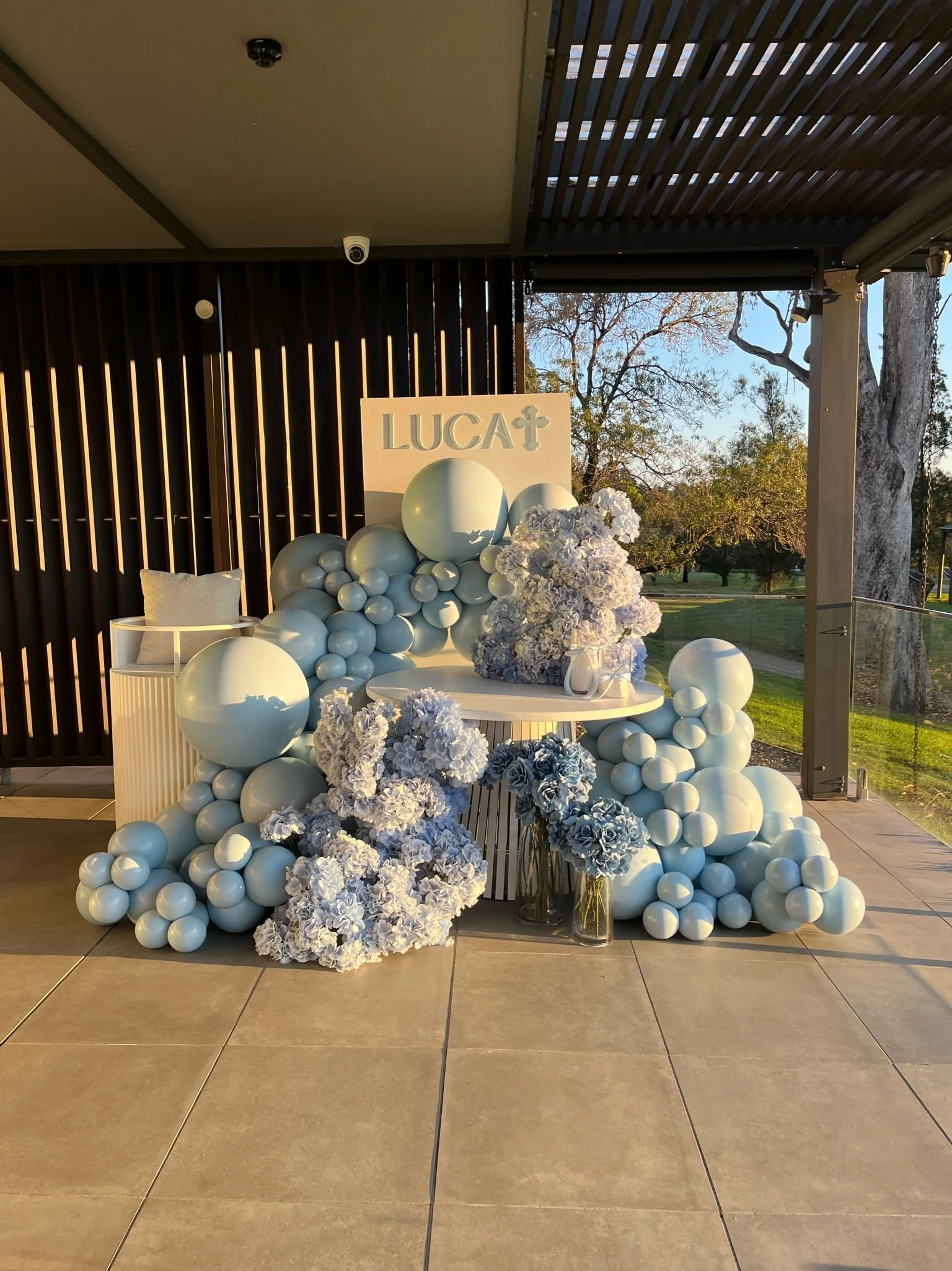 Decorative display with white and light blue balloons, white floral arrangements, and a sign reading "LUCA" with a cross symbol, set outdoors during sunset.