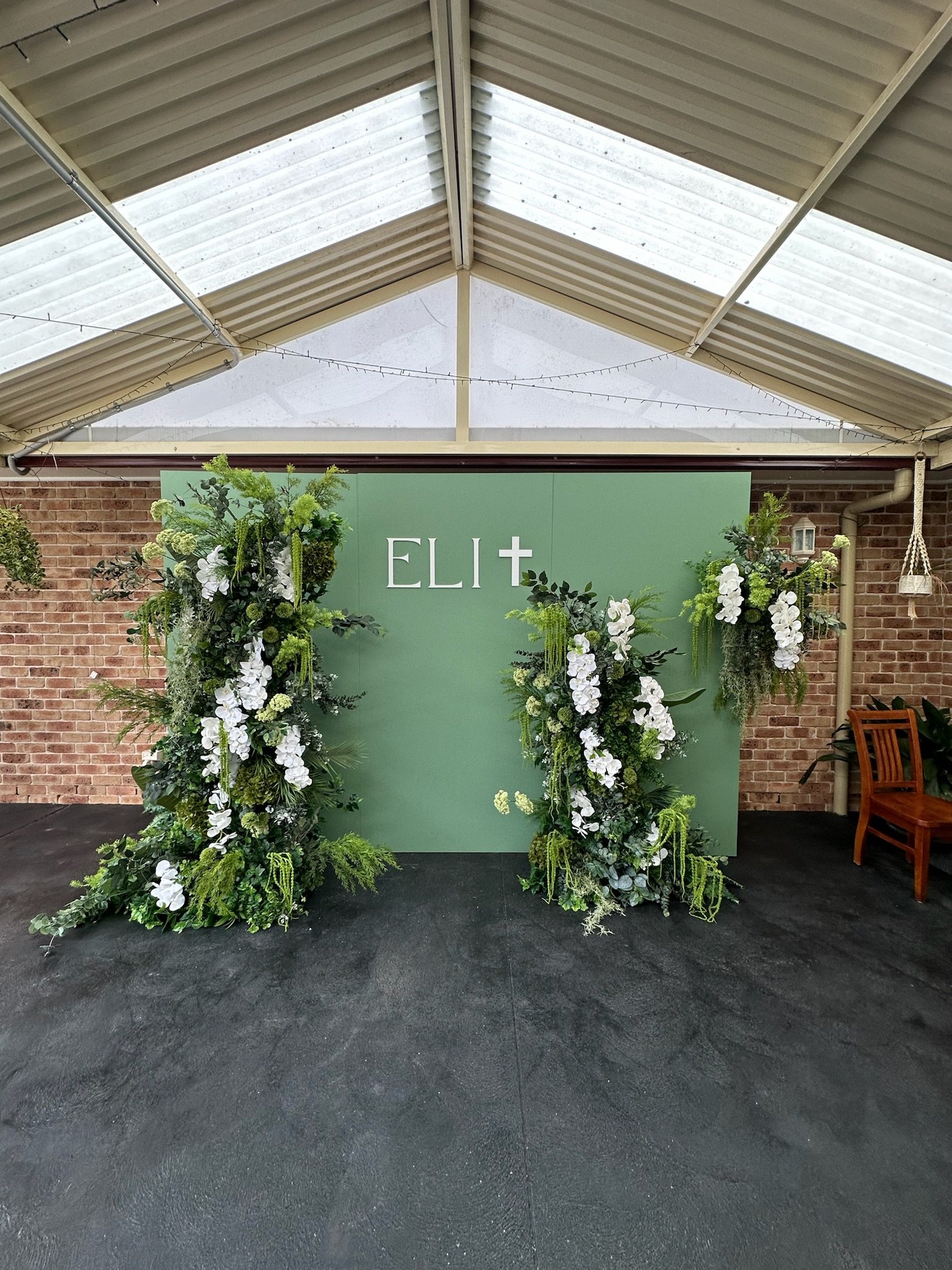 Decorative green backdrop with white lettering and cross, flanked by large floral arrangements of white flowers and green foliage, set inside a covered venue with brick walls.