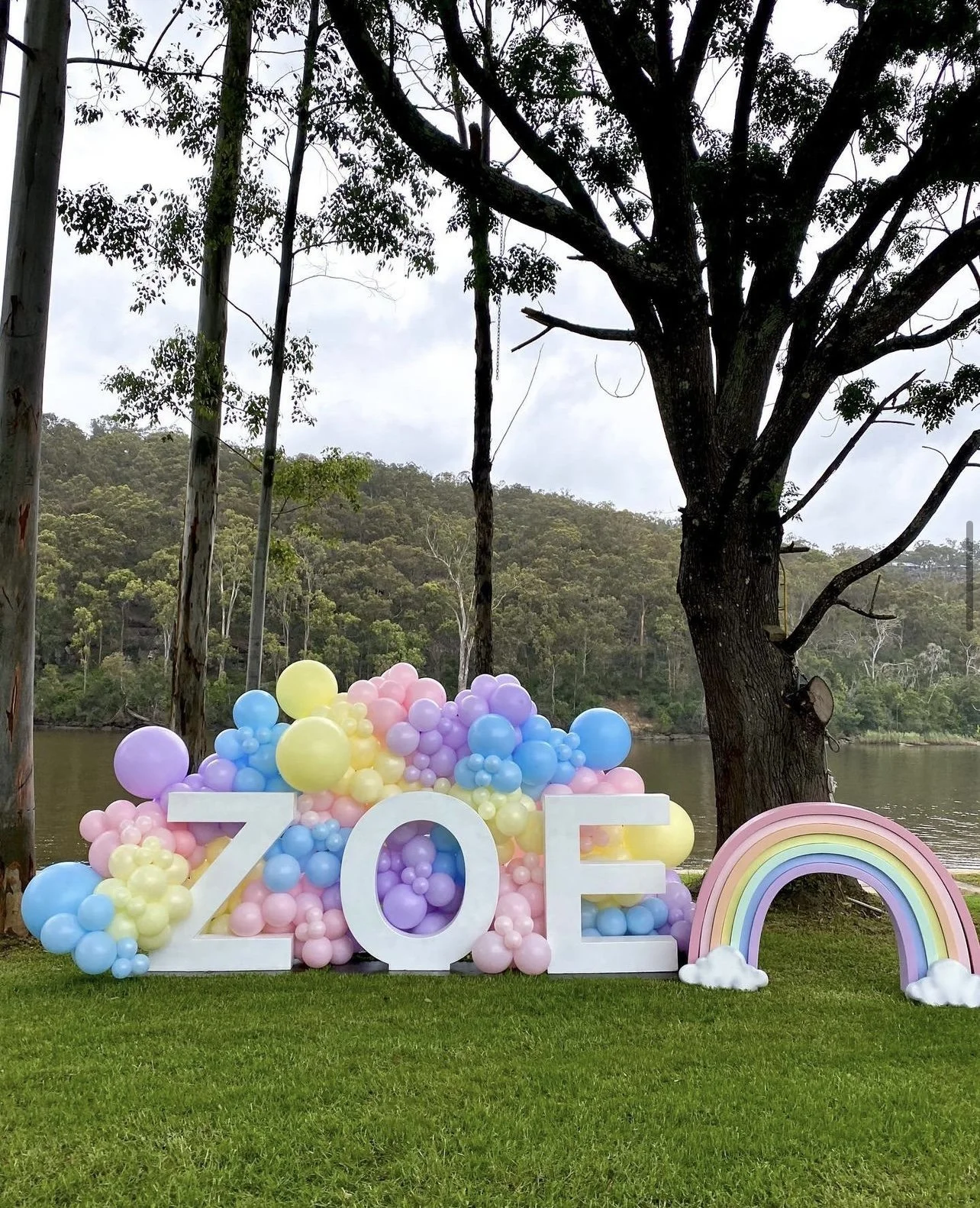 Colorful balloon display spelling out 'ZOE' near a rainbow decoration, set on grass beside a large tree and a river with hilly wooded landscape in the background.