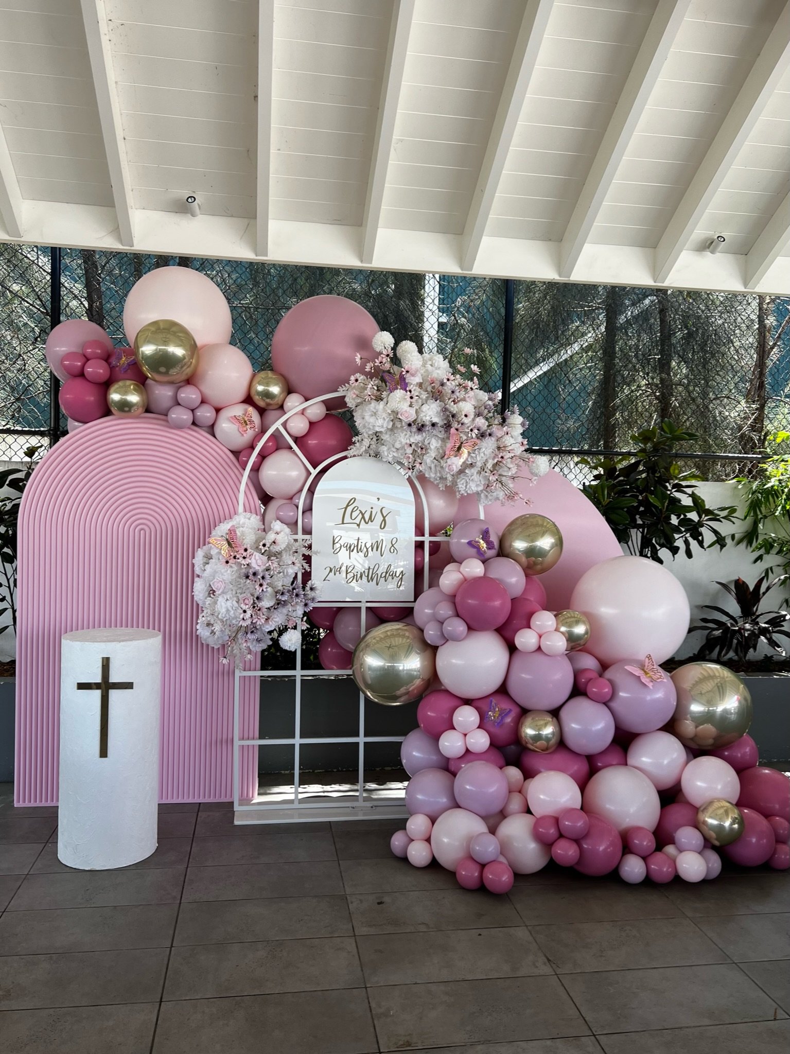 Decorative balloon display for a baptism and 2nd birthday celebration with pink, purple, white, and gold balloons, floral arrangements, a sign with the name "Lexis," and a white pillar with a cross.