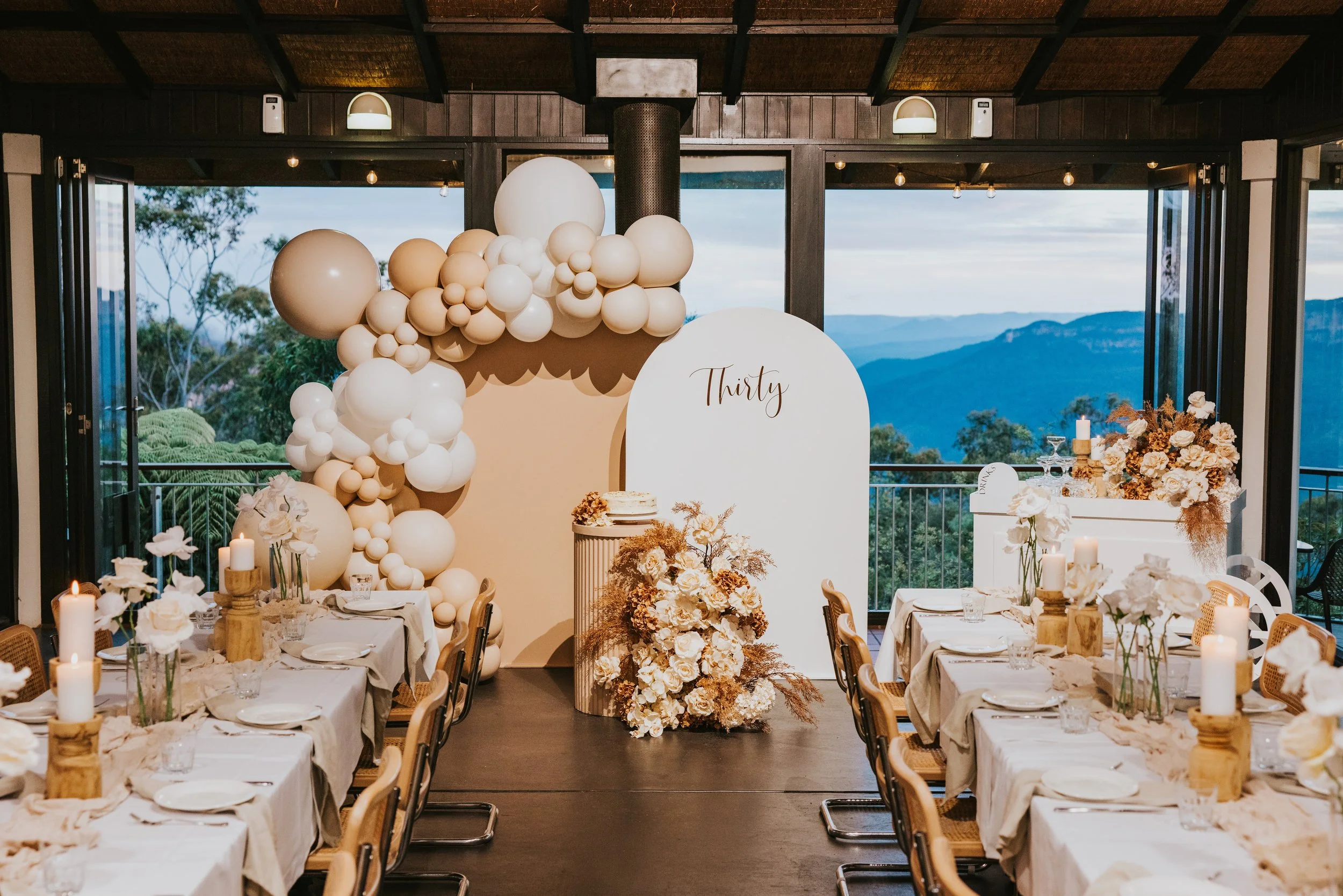 Elegant indoor celebration space with long tables decorated with white flowers, candles, and neutral-colored tablecloths. A backdrop with a balloon arrangement in shades of beige and white, and a floral sign with the number 'Thirty'. Large windows re