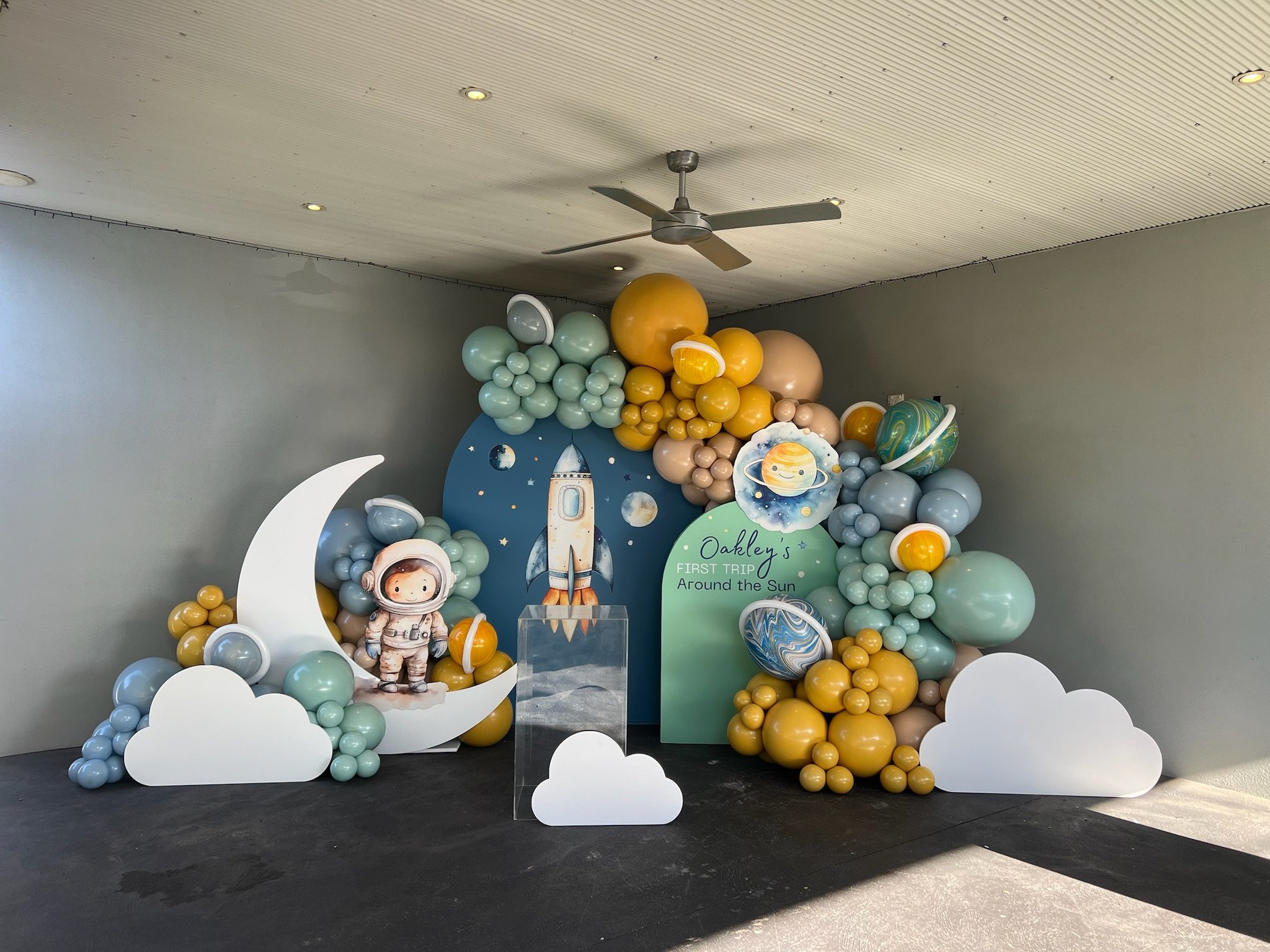 Space-themed birthday decoration with balloons, clouds, a moon, planets, and a rocket, celebrating Oakley's first trip around the sun.