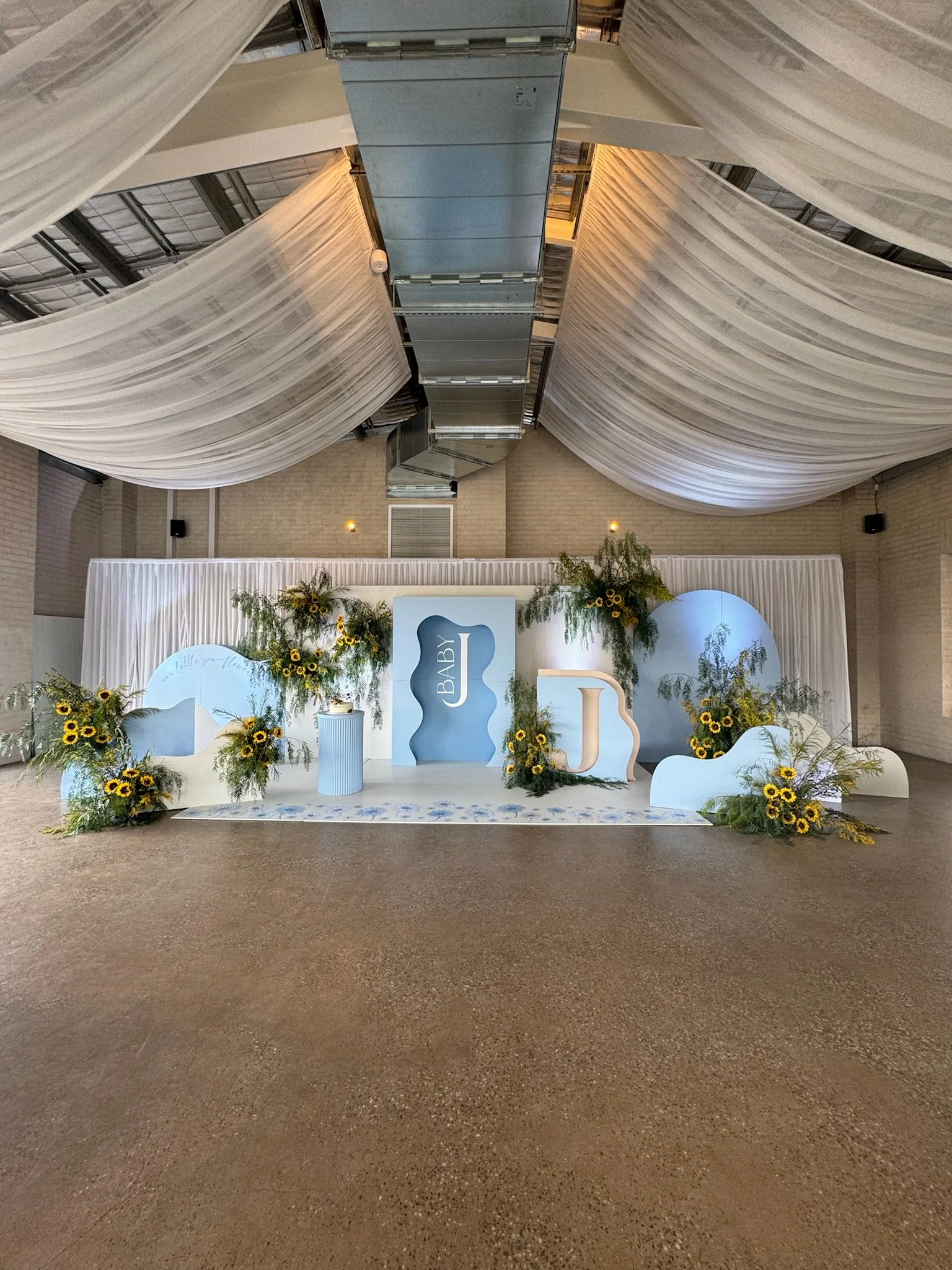 Decorative baby shower setup with blue and white accents, floral arrangements featuring sunflowers, and large stylized letters spelling out 'BABY' with the letter 'J'.
