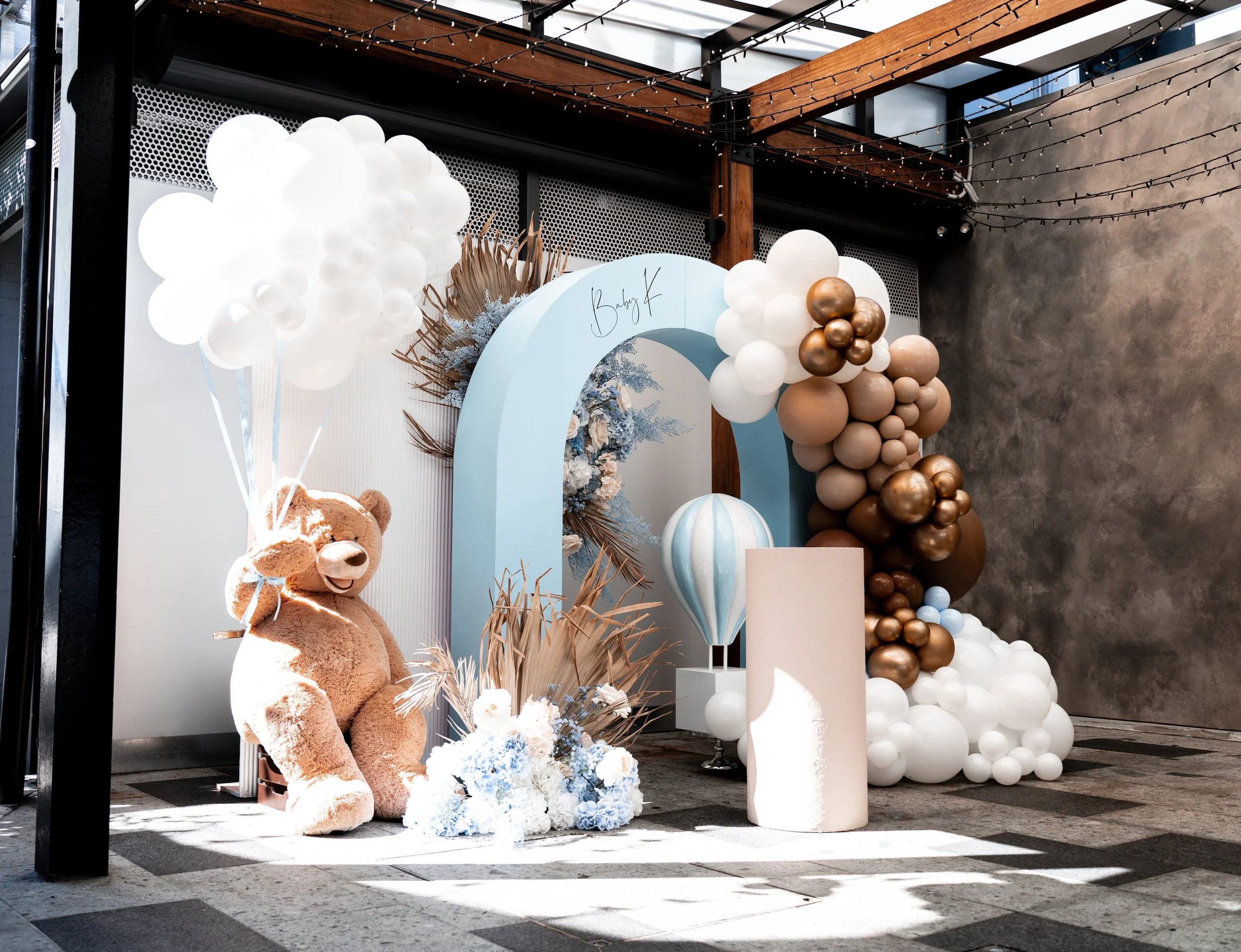 Decorative backdrop for a baby shower with balloons, a teddy bear holding balloon bundles, a large blue arch with the words "Baby Girl," floral arrangements, and a striped hot air balloon prop.