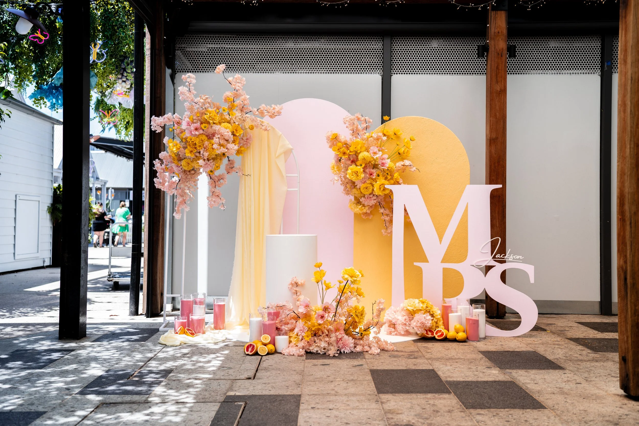 Decorative display with pink and yellow flowers, large letter 'M' and 'Y' and some candles and fruit in front, set outdoors under a covered area.