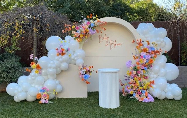 Decorative backdrop with white balloons and colorful flowers on a grassy lawn, featuring a white cylindrical table in front and handwritten 'Baby on Board' sign.