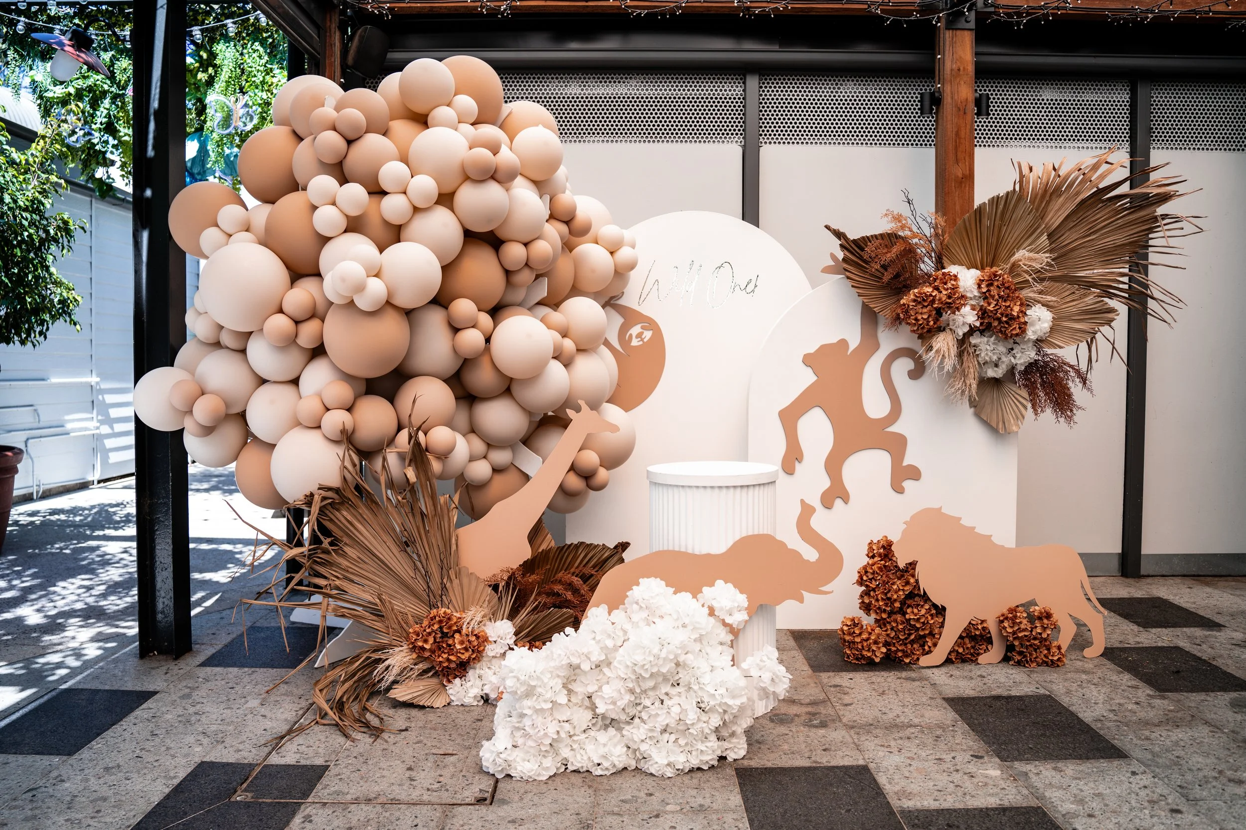 Decorative display with a large cluster of beige and white balloons, cutouts of animals such as an elephant, lion, and flamingo, along with dried and artificial flowers, set in a semi-outdoor space with string lights.
