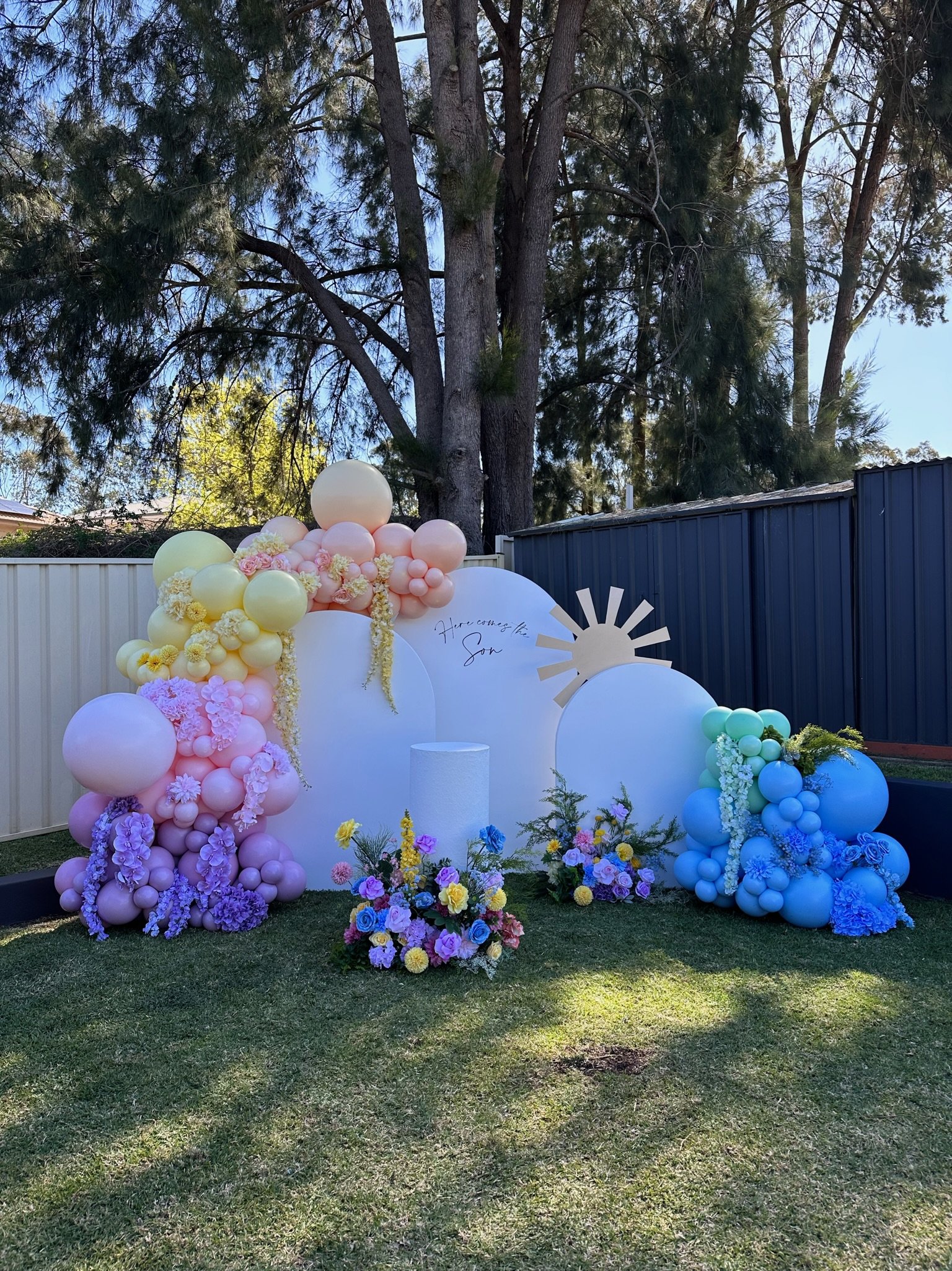 Decorative backdrop with pastel-colored balloons, flowers, and a sun cutout, set outdoors in a backyard with trees and fencing.