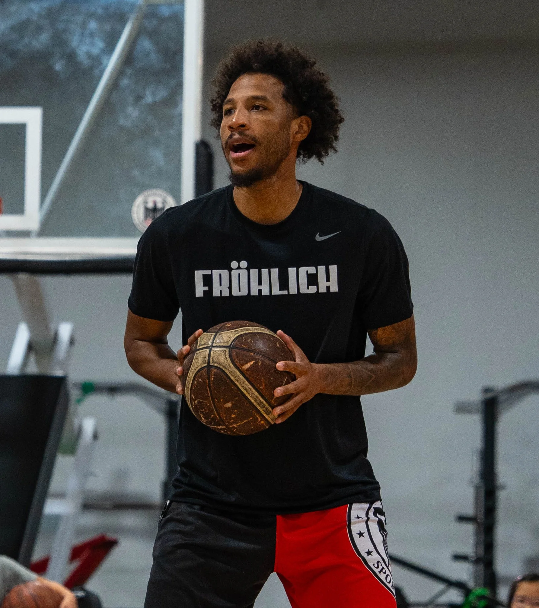 A man playing basketball indoors, holding a basketball with both hands, wearing a black shirt with 'FRÖHLICH' printed on it and red and black shorts.