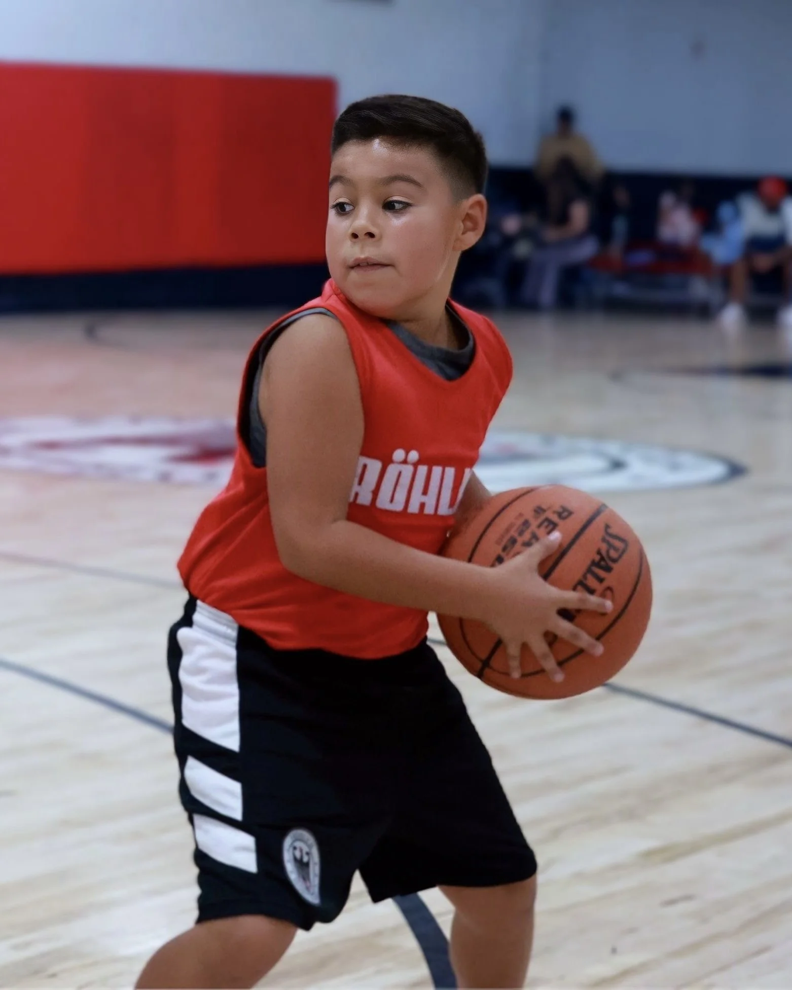 Young boy playing basketball on court, holding basketball, wearing red and black sports uniform.
