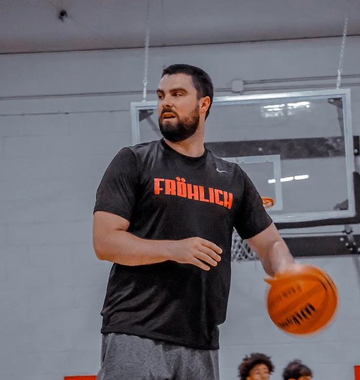 A man with a beard playing basketball in an indoor gym, wearing a black T-shirt with red letters that say 'FROHLICH' and gray shorts.