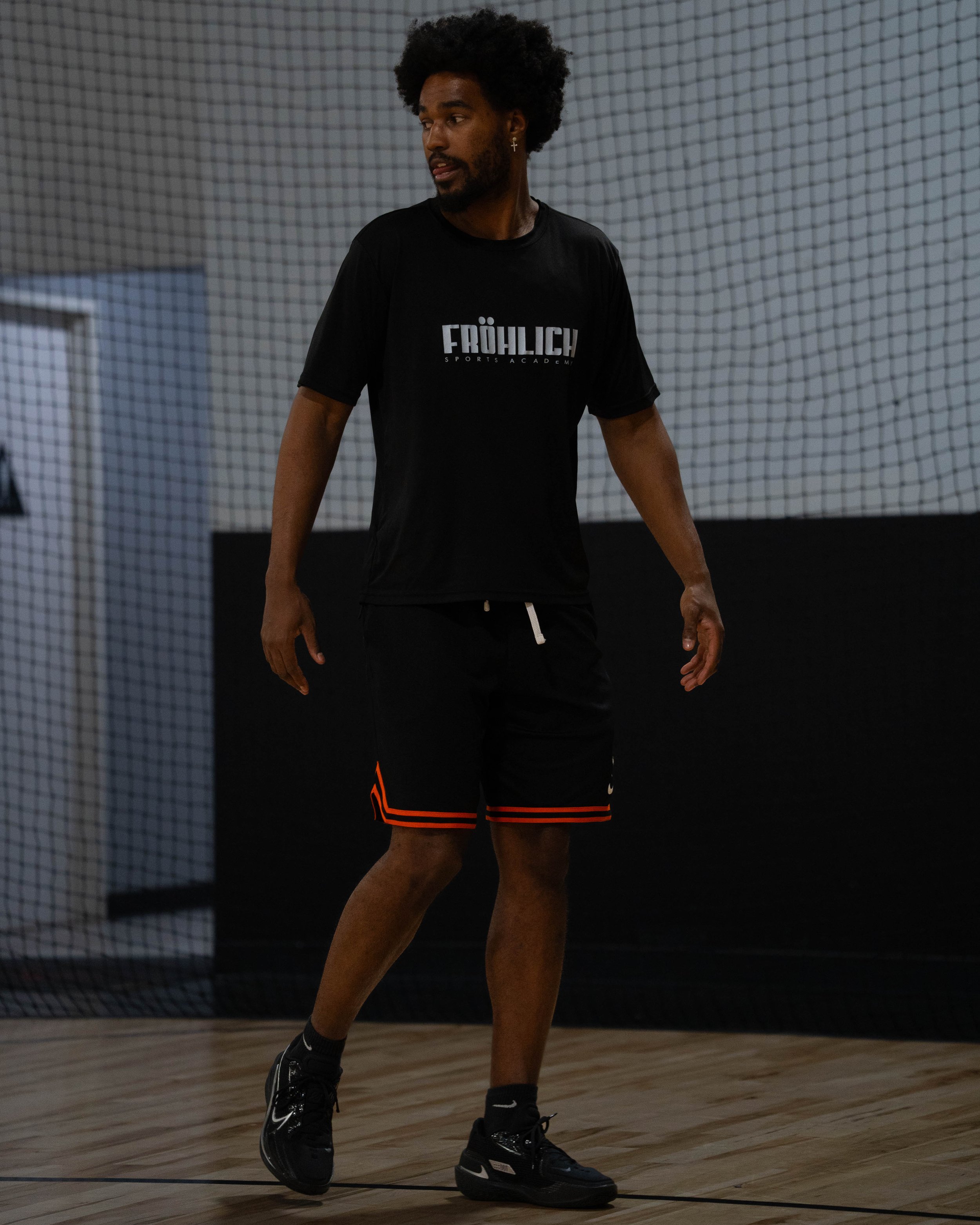 A man with curly hair and a beard wearing a black T-shirt with 'FRÖHLICH SPORTS ACADEMY' written on it, black shorts with red trim, and black shoes standing on a wooden indoor court.