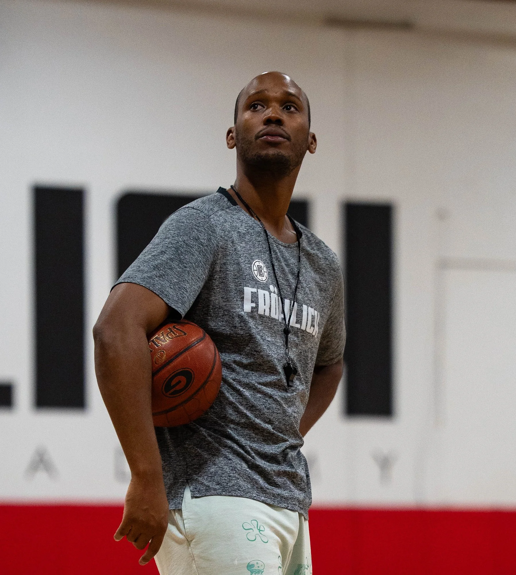 A man standing indoors holding a basketball under his arm, wearing a gray T-shirt with 'FROLIC' on it and light-colored shorts with green clover and mushroom designs.