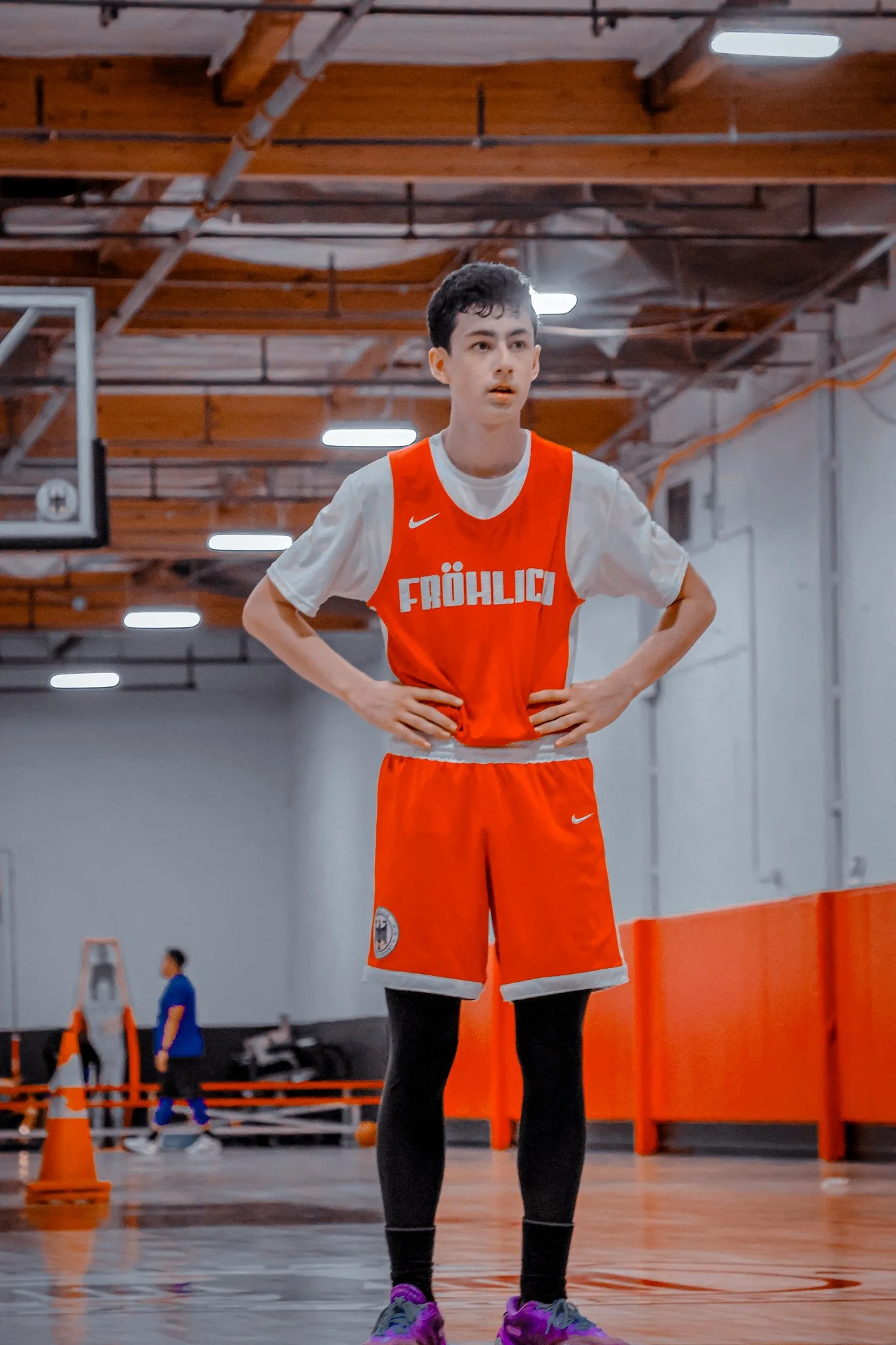 A young male basketball player wearing a red and white jersey with 'FÖHLIG' on the front, red shorts, purple sneakers, and black tights, standing on an indoor basketball court with hands on hips, looking focused.