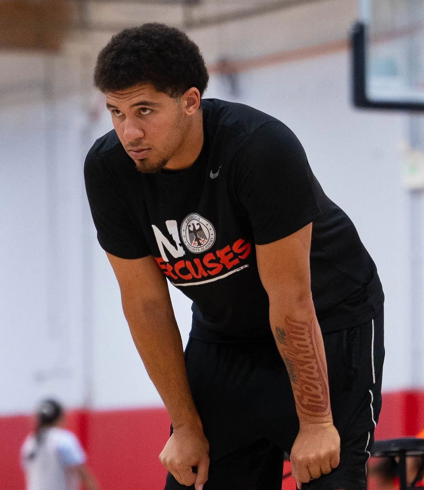 Young man in black athletic clothing with a tattoo on his left arm, leaning forward with hands on his knees in an indoor sports facility.