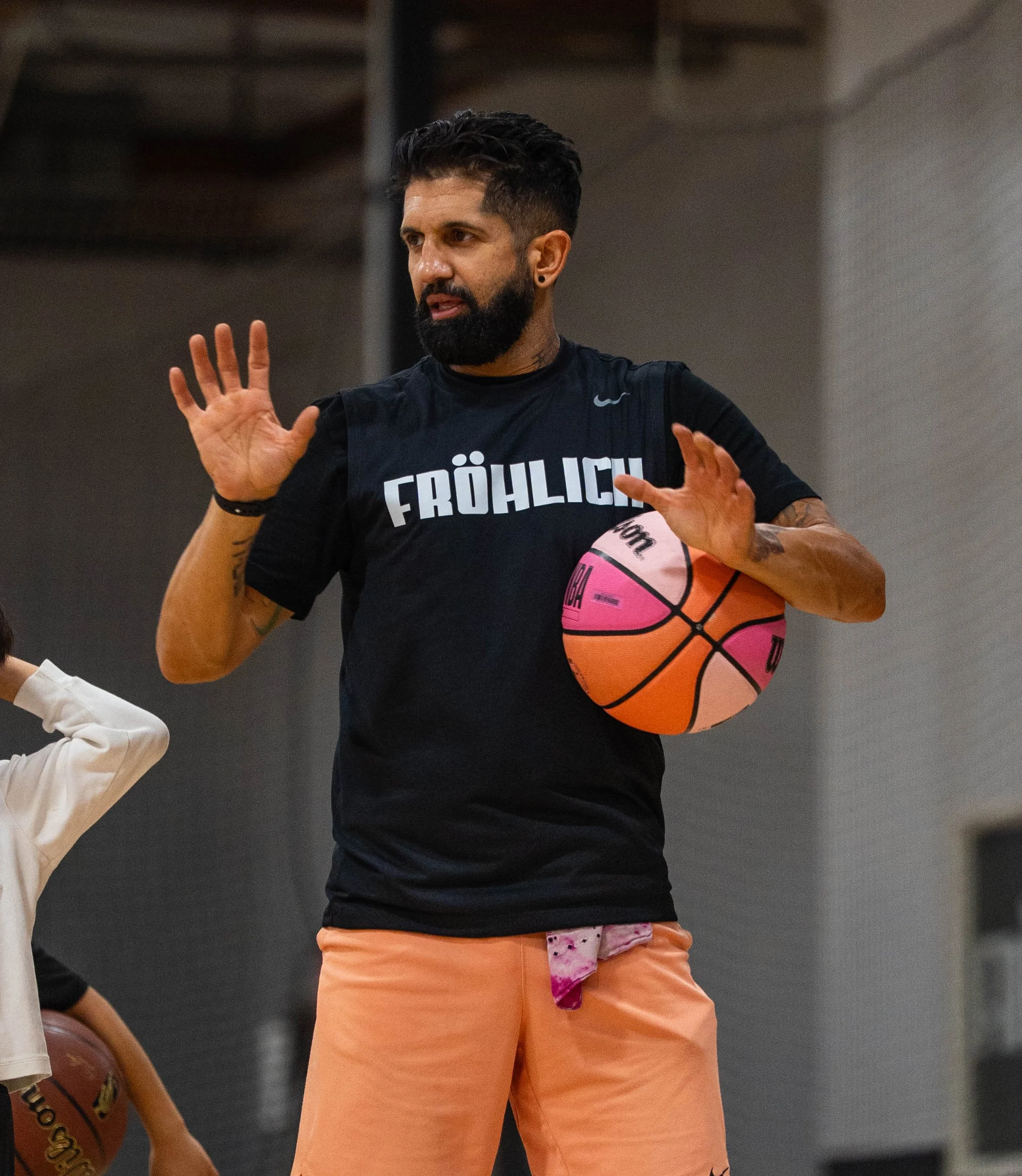 A man with a beard and tattoos, wearing a black T-shirt with the word 'FRÖHLICH' and peach-colored shorts, holding a pink and orange basketball under his right arm, standing indoors in a gym or sports facility.