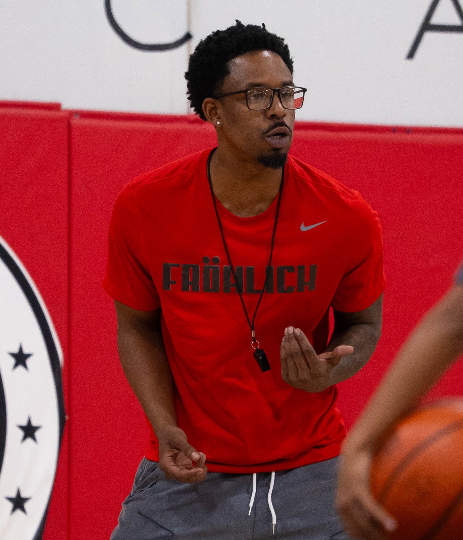 A man wearing glasses and a red t-shirt with the word 'FRÖHLICH' printed on it, holding a basketball and instructing during a practice or game in an indoor gym with red padded walls.
