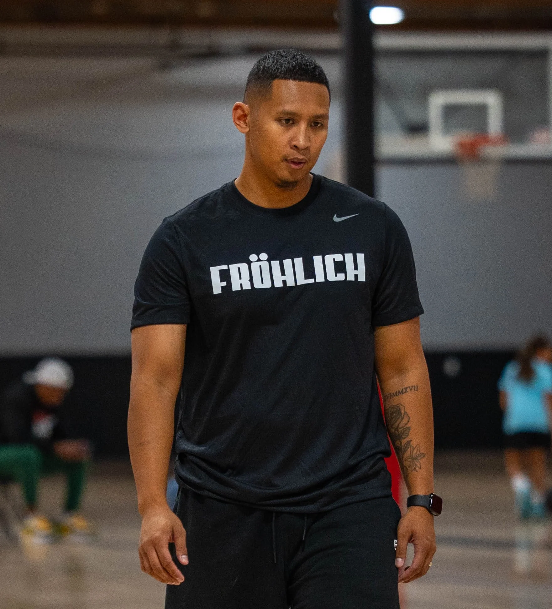A young man wearing a black shirt with the word 'FRÖHLICH' on it, standing in an indoor basketball court.