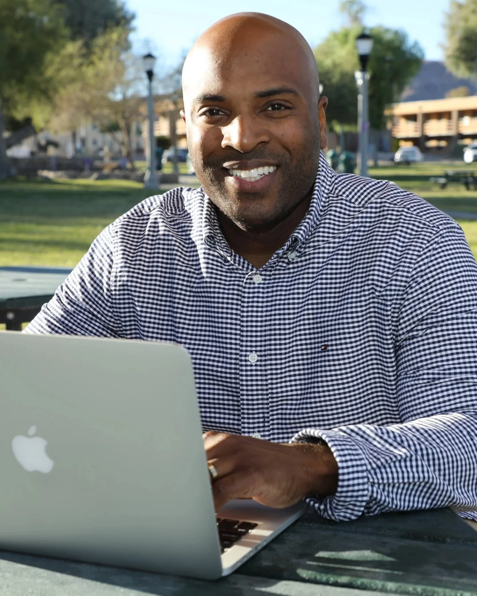 A man smiling while sitting outdoors at a table with a silver MacBook laptop in front of him.