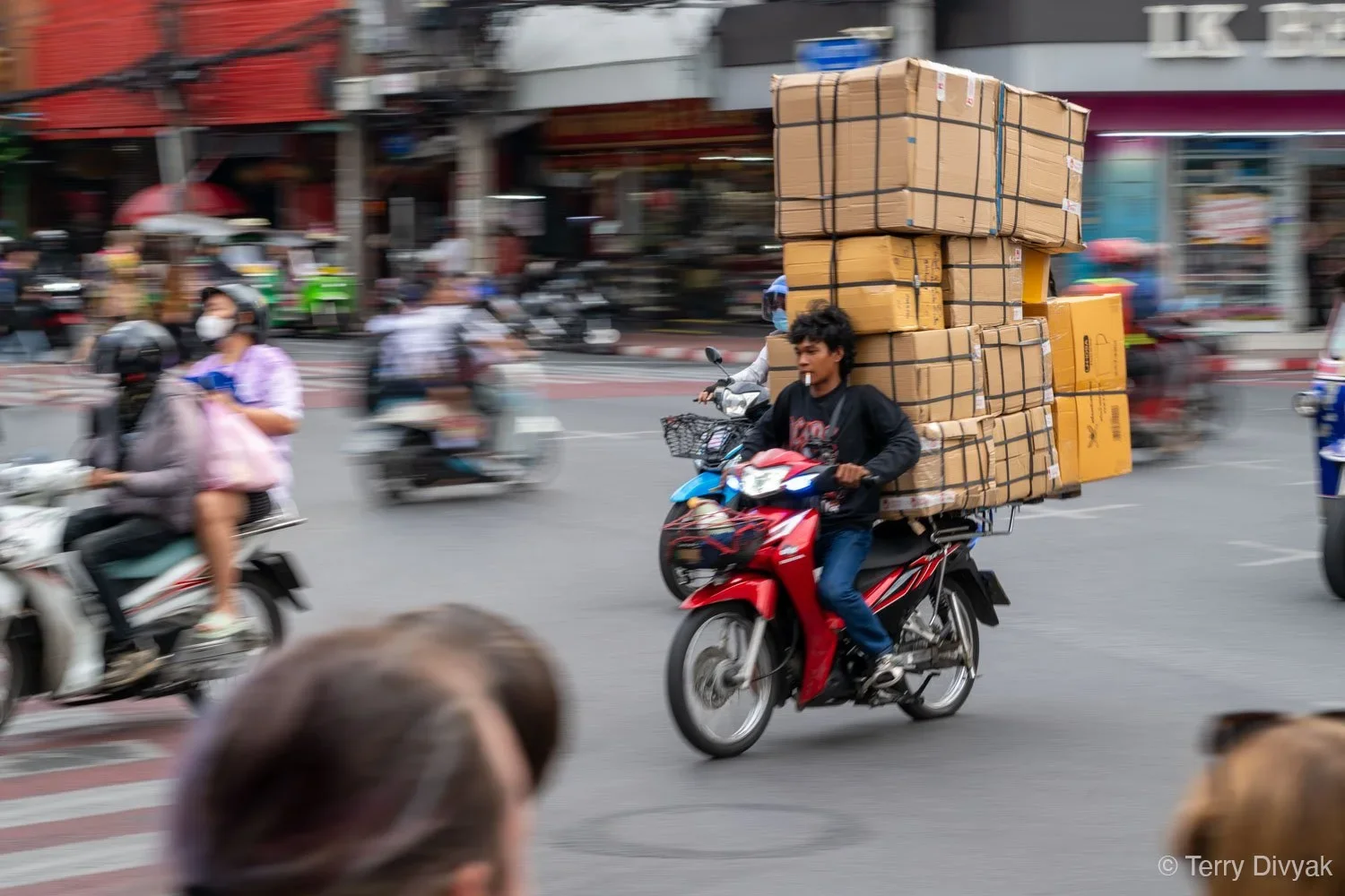 A man riding a red motorcycle on a busy city street, carrying large boxes of cardboard boxes stacked on his back and secured with straps.