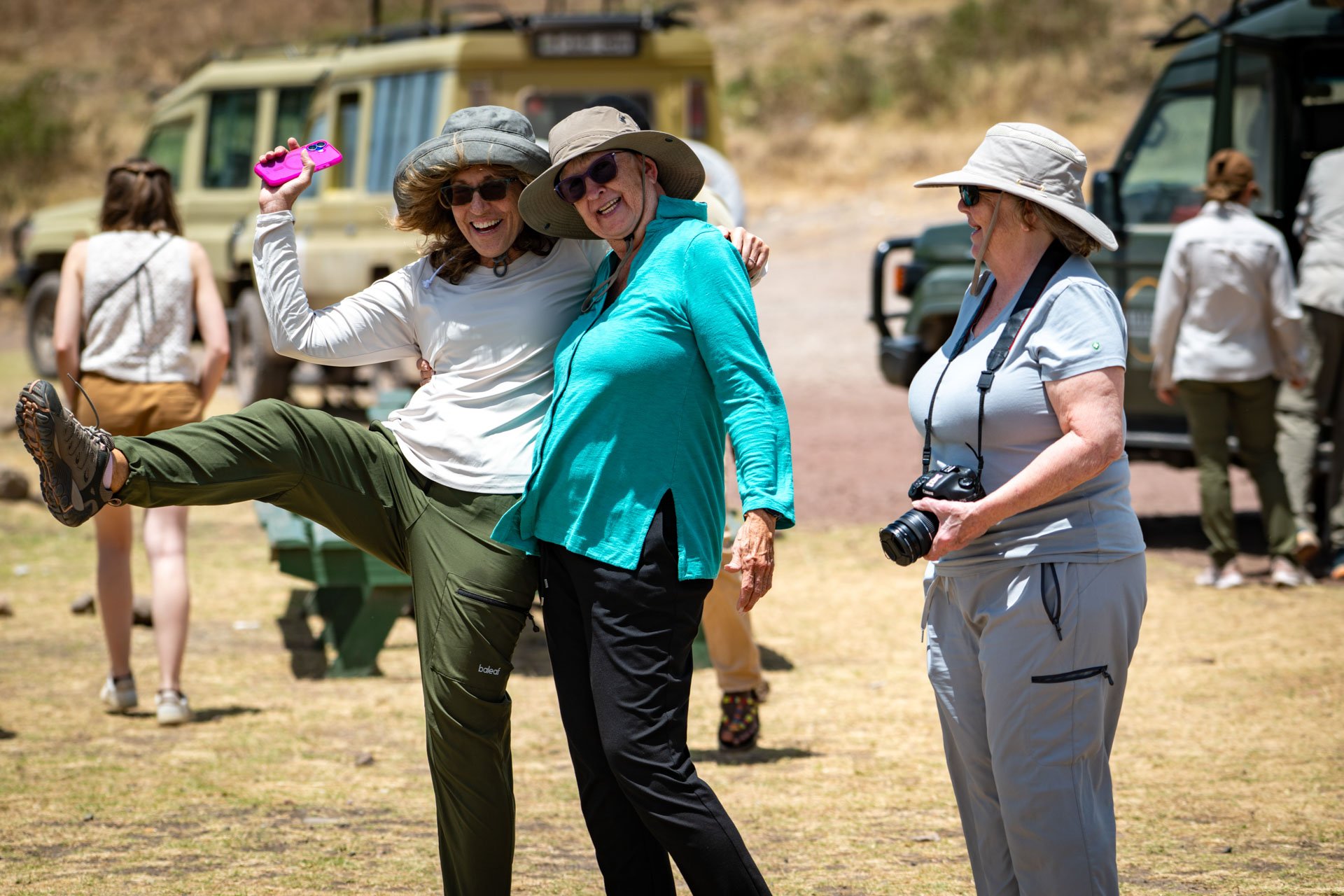 Group of women outdoors, smiling and having fun, with some dressed in hiking or safari attire, near vehicles, in a sunny, dry outdoor setting.