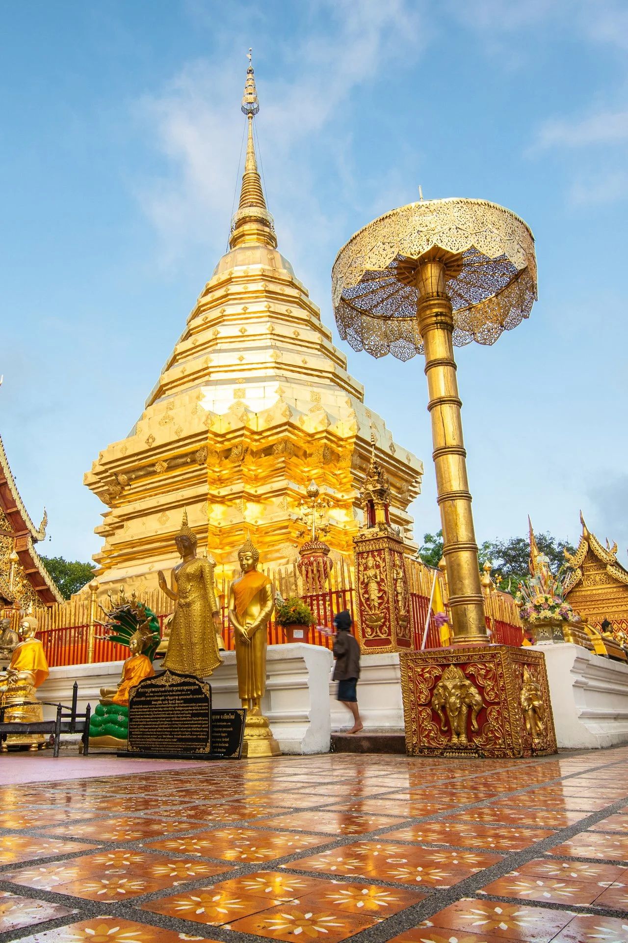 A golden pagoda with a tiered spire, surrounded by golden statues and an ornate parasol, set against a blue sky in a temple courtyard.