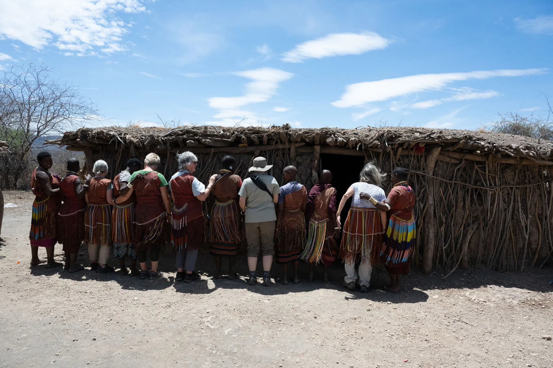 Group of people, some in traditional Maasai attire, standing and looking into a thatched hut in a rural setting with dry ground and sparse trees, under a blue sky with clouds.