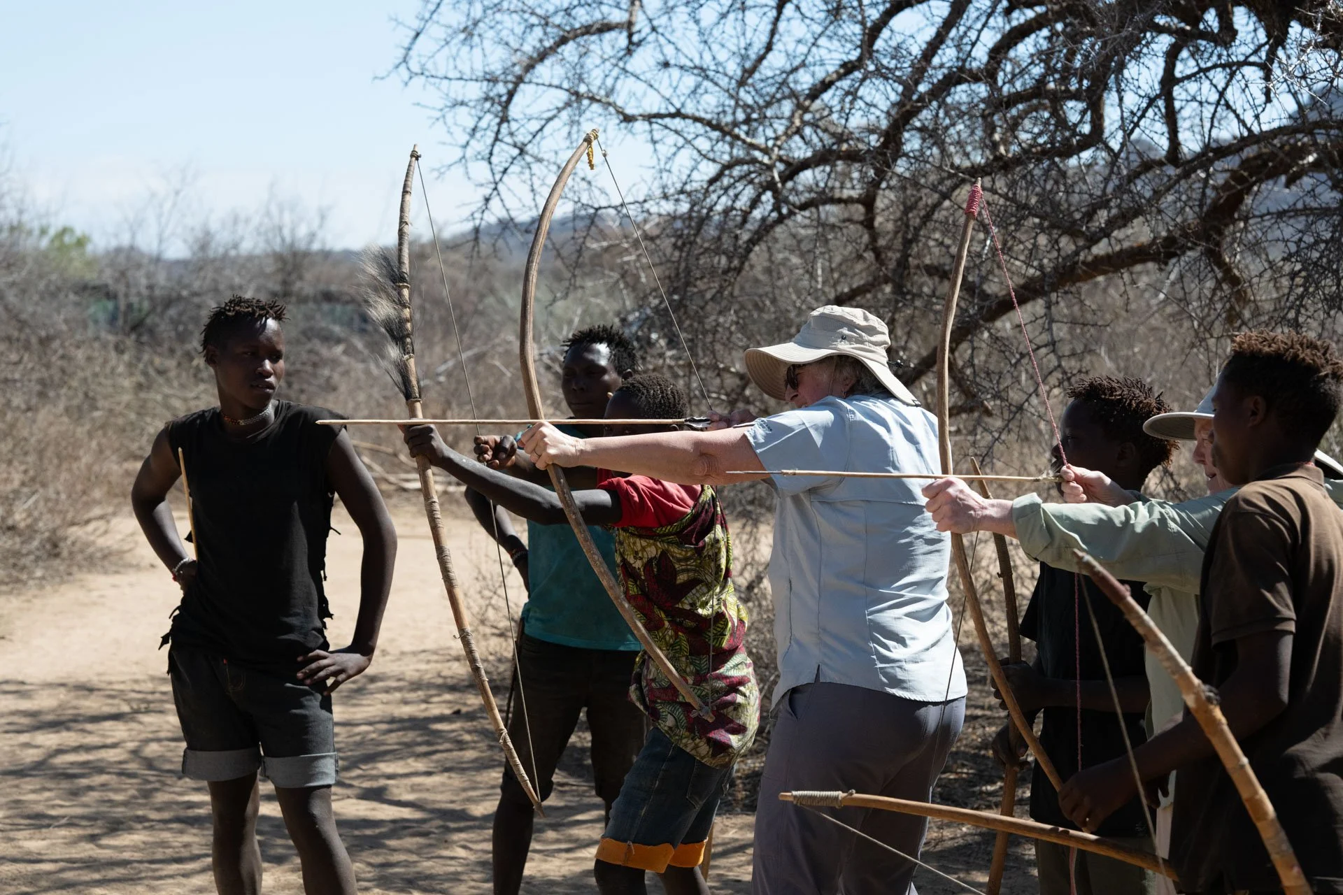 A group of young Hadzabe people and an adult with a hat practicing archery outdoors in a dry, wooded area.