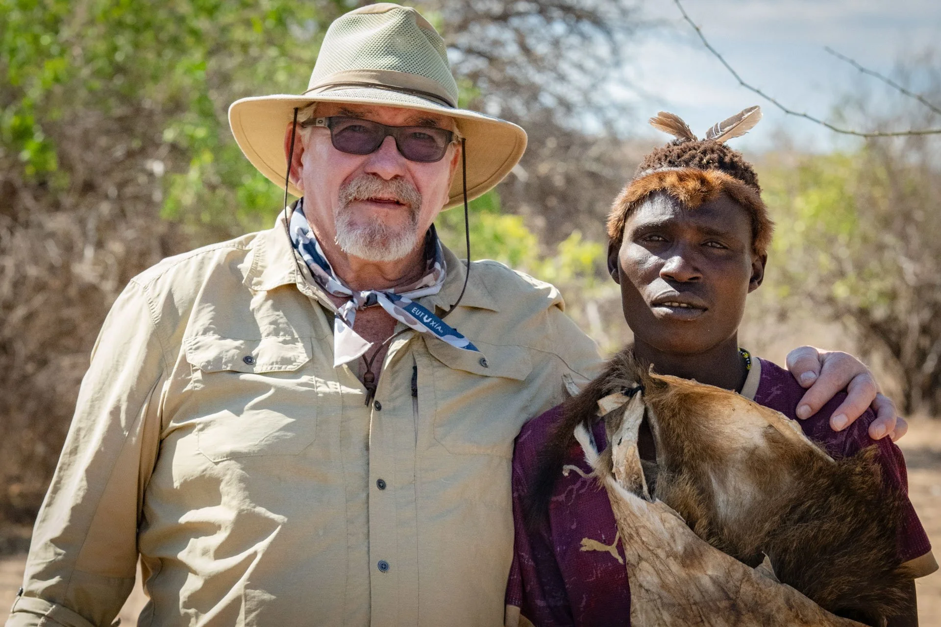An older man with a beard, wearing a wide-brimmed hat, sunglasses, and light-colored shirt, standing next to a young Hadzabe man in Tanzania, with dark skin and short hair, dressed in traditional clothing with feathers and animal skin. 