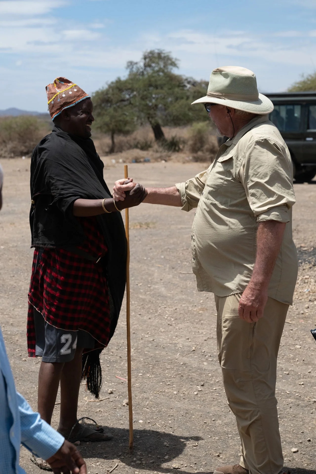 A Datoga man in traditional clothing shaking hands with a man in beige safari clothing in a desert landscape with sparse trees.