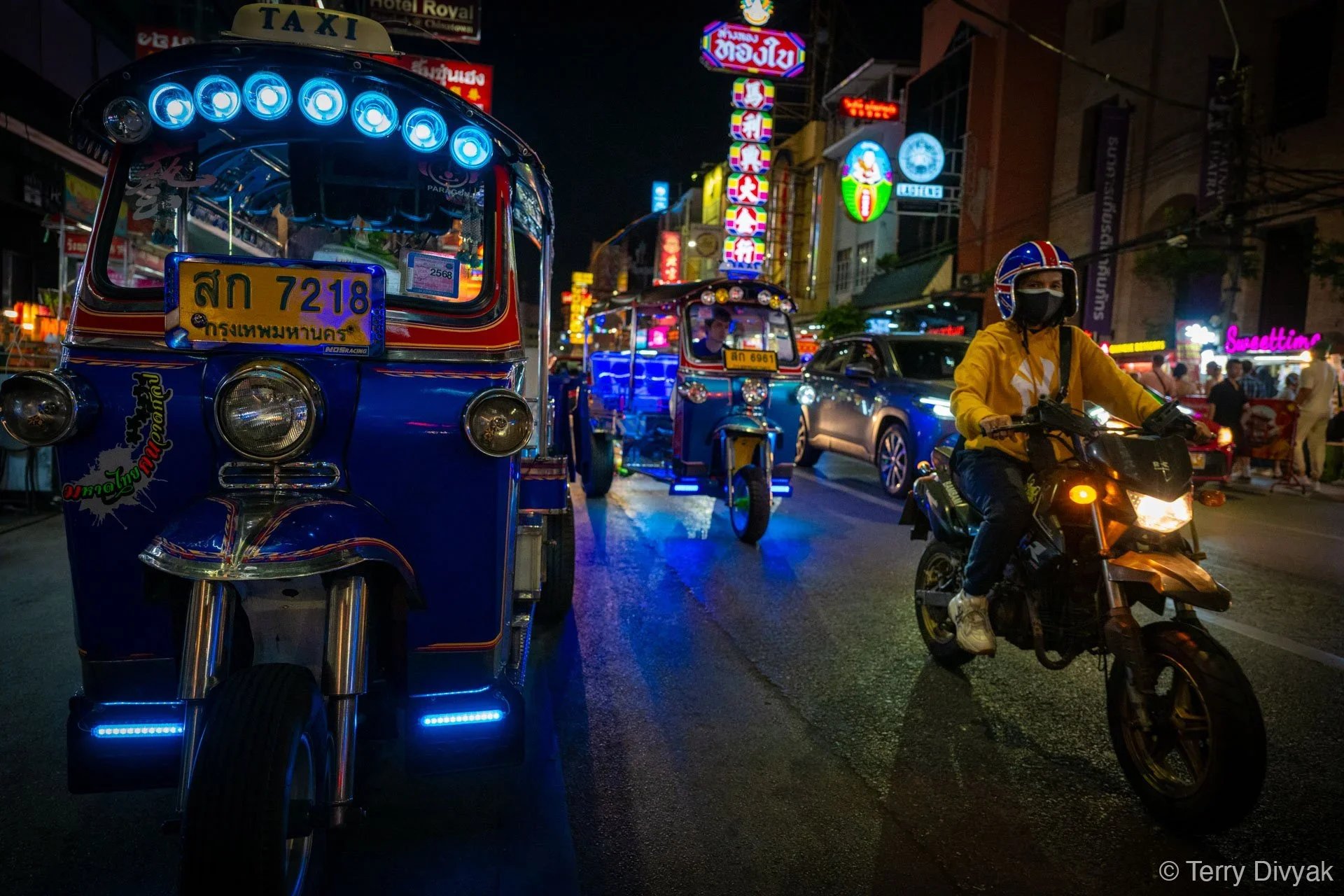 Night scene of a busy street in Thailand with colorful neon signs, a blue tuk-tuk, a motorcycle rider wearing a yellow jacket and black mask, and other vehicles and pedestrians.