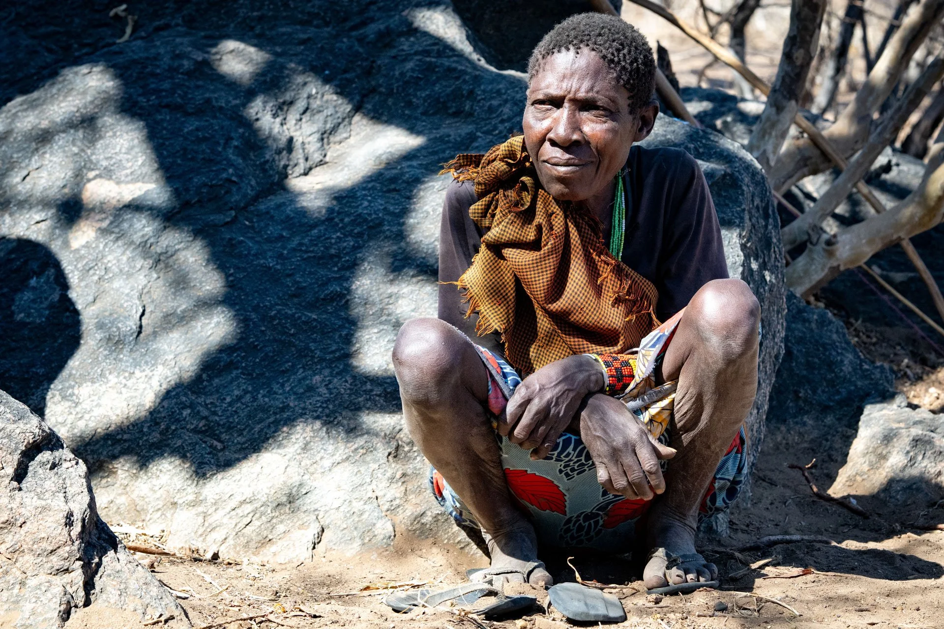 An elderly person with dark skin, short curly hair, wearing a dark shirt, colorful patterned shorts, and a yellow scarf, sitting on the ground amidst rocks and dirt. They are looking off into the distance with a reflective expression, with some trees