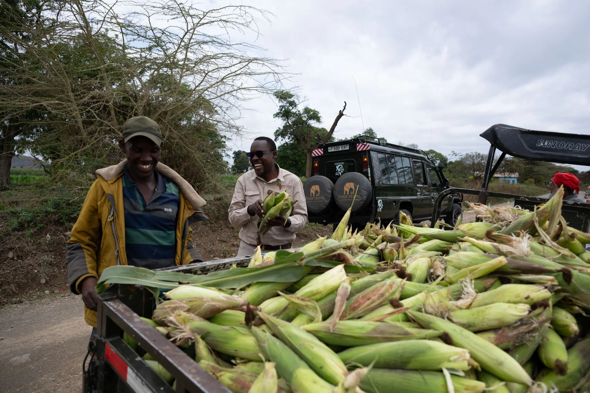 Two men smiling near a trailer loaded with freshly harvested corn cobs, with a black safari vehicle and a person wearing a red headscarf in the background, outdoors on a cloudy day.