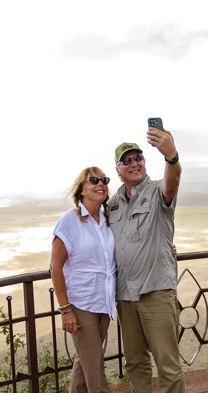 A smiling couple taking a selfie outdoors against a cloudy sky and landscape. The woman wears sunglasses, a white shirt, and tan pants, while the man wears sunglasses, a green cap, a gray shirt, and tan pants.