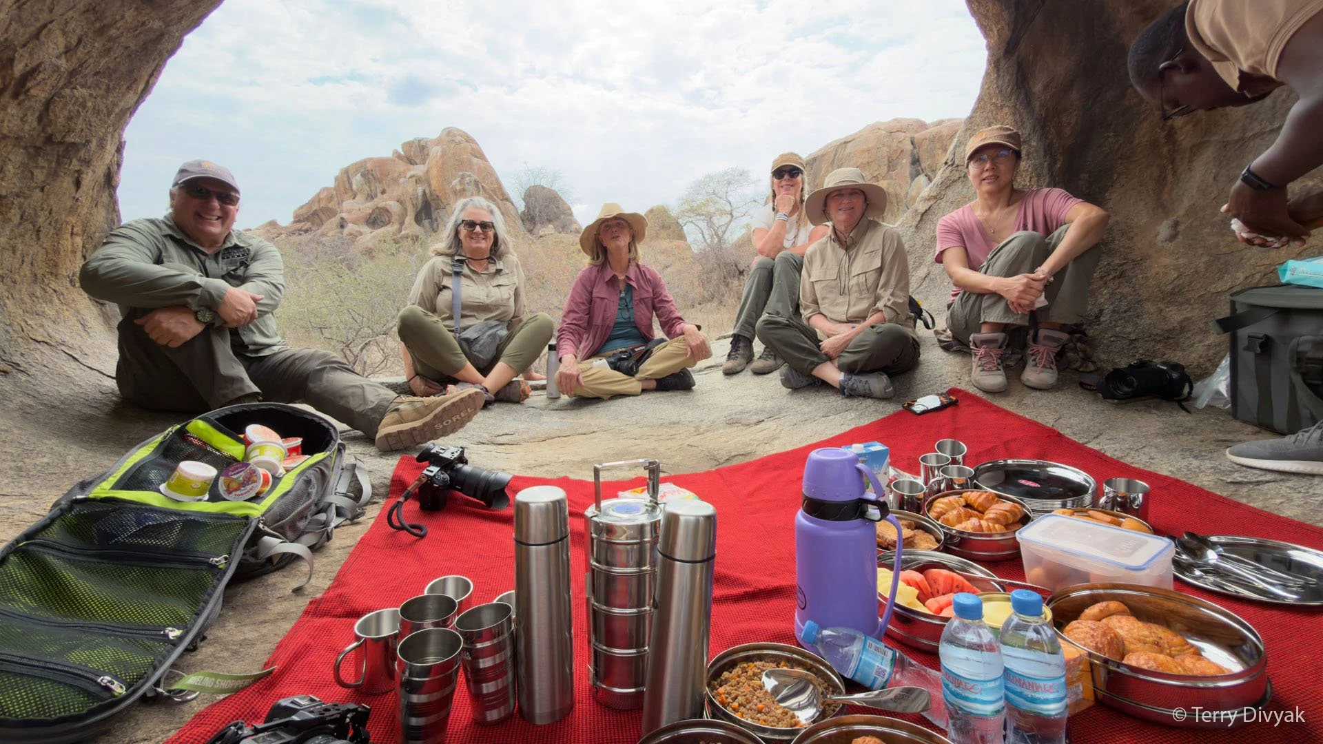 A group of people sitting on the ground in a desert landscape with rocky formations, gathered for a picnic under natural rock formations, with food and beverages on a red blanket in the foreground.