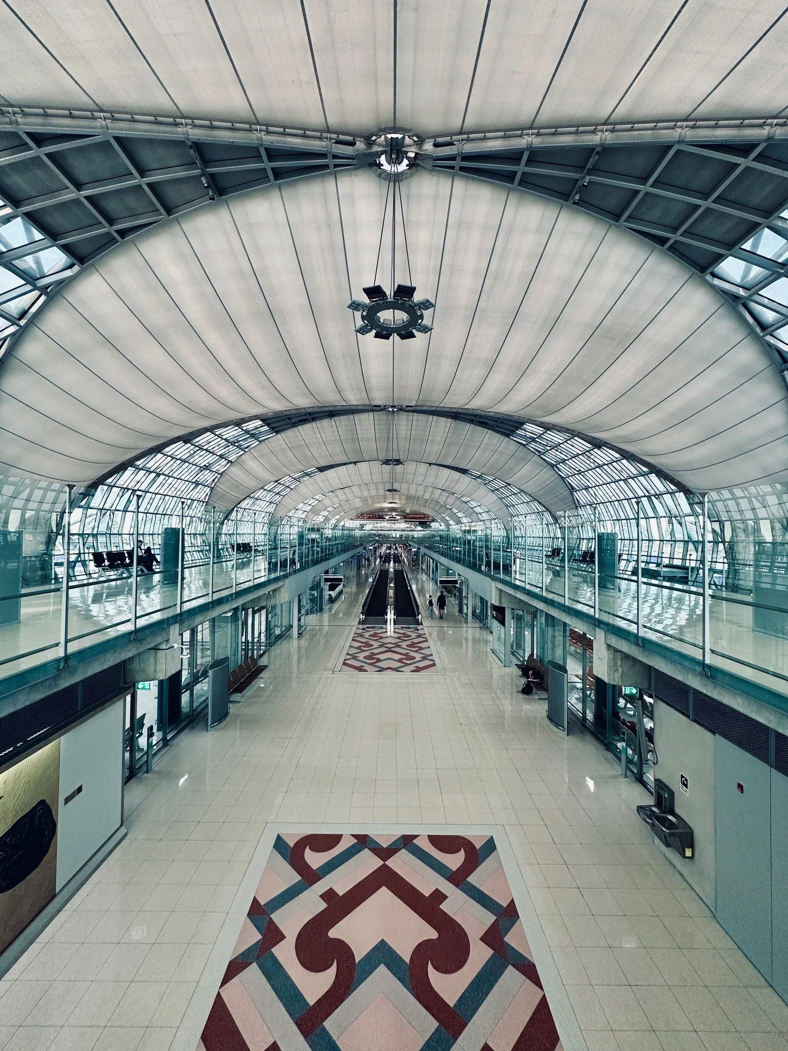 Interior of a modern airport terminal with glass walls, an arched ceiling, and a long corridor with patterned floor tiles.