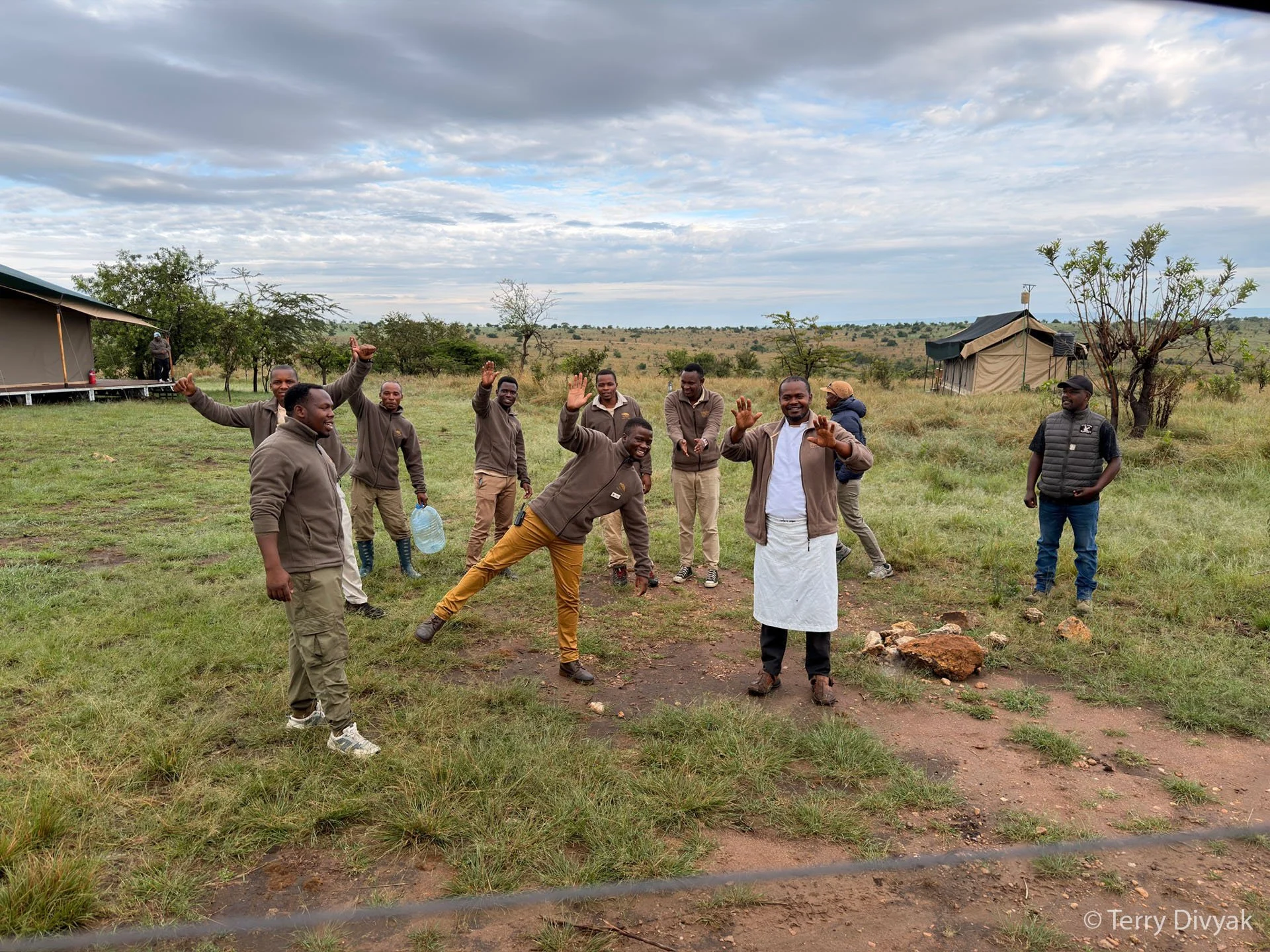 Group of men and boys standing outdoors in a grassy area, smiling and waving at the camera, with some tents and trees in the background under a cloudy sky.