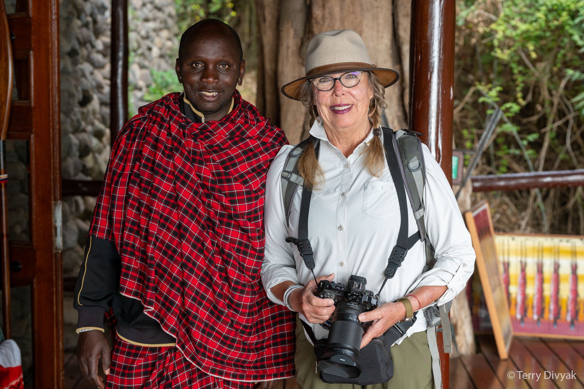 Two women standing together outdoors, one in traditional Maasai clothing and the other in casual hiking attire, holding a camera. They are smiling.