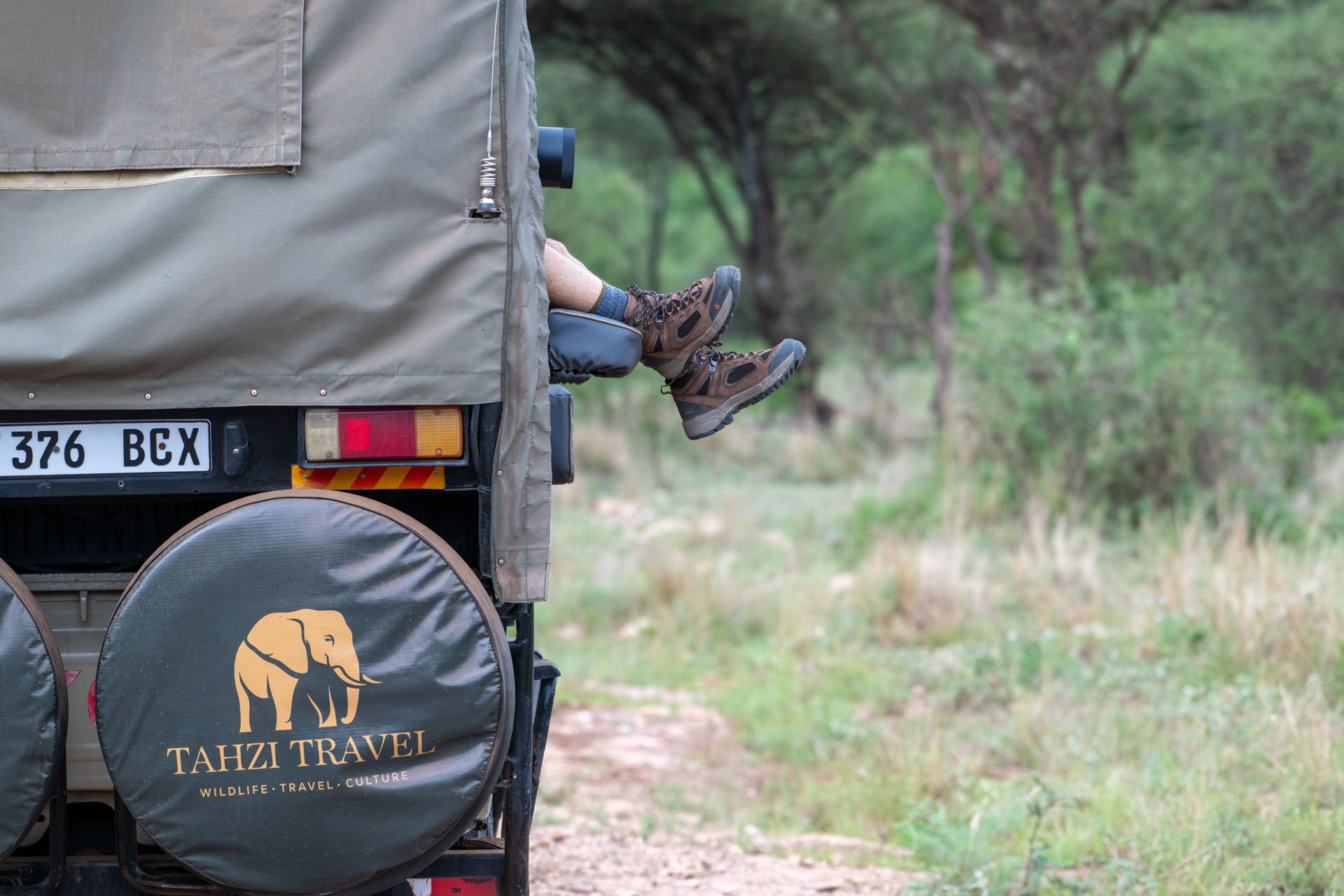 Back of a Tahzi Travel safari vehicle with two pairs of feet hanging out of a window, surrounded by a grassy and wooded landscape.