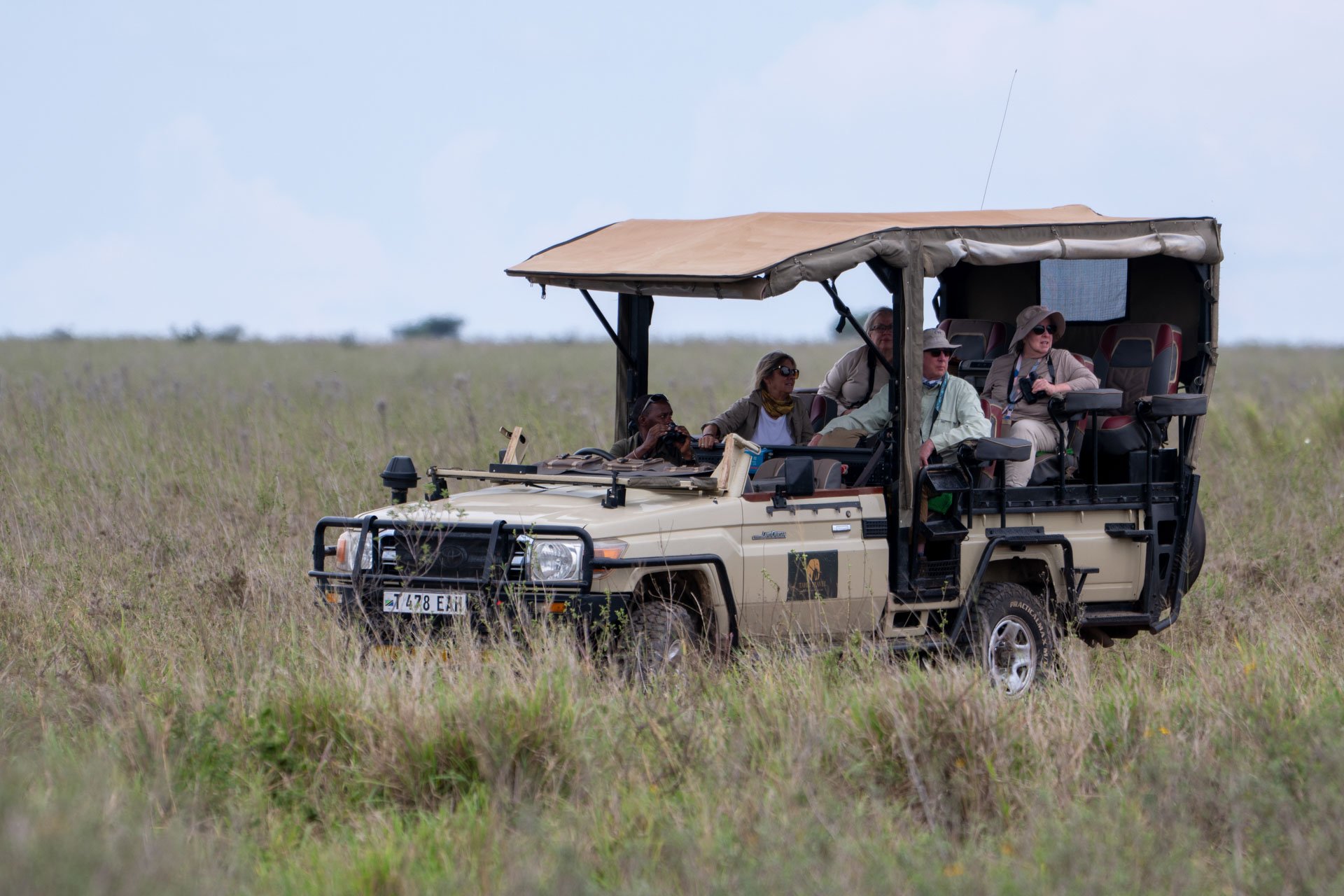 A group of six tourists on a safari tour in an open-air vehicle with Tahzi Travel in a grassy plain, with some of them taking photos and observing wildlife.
