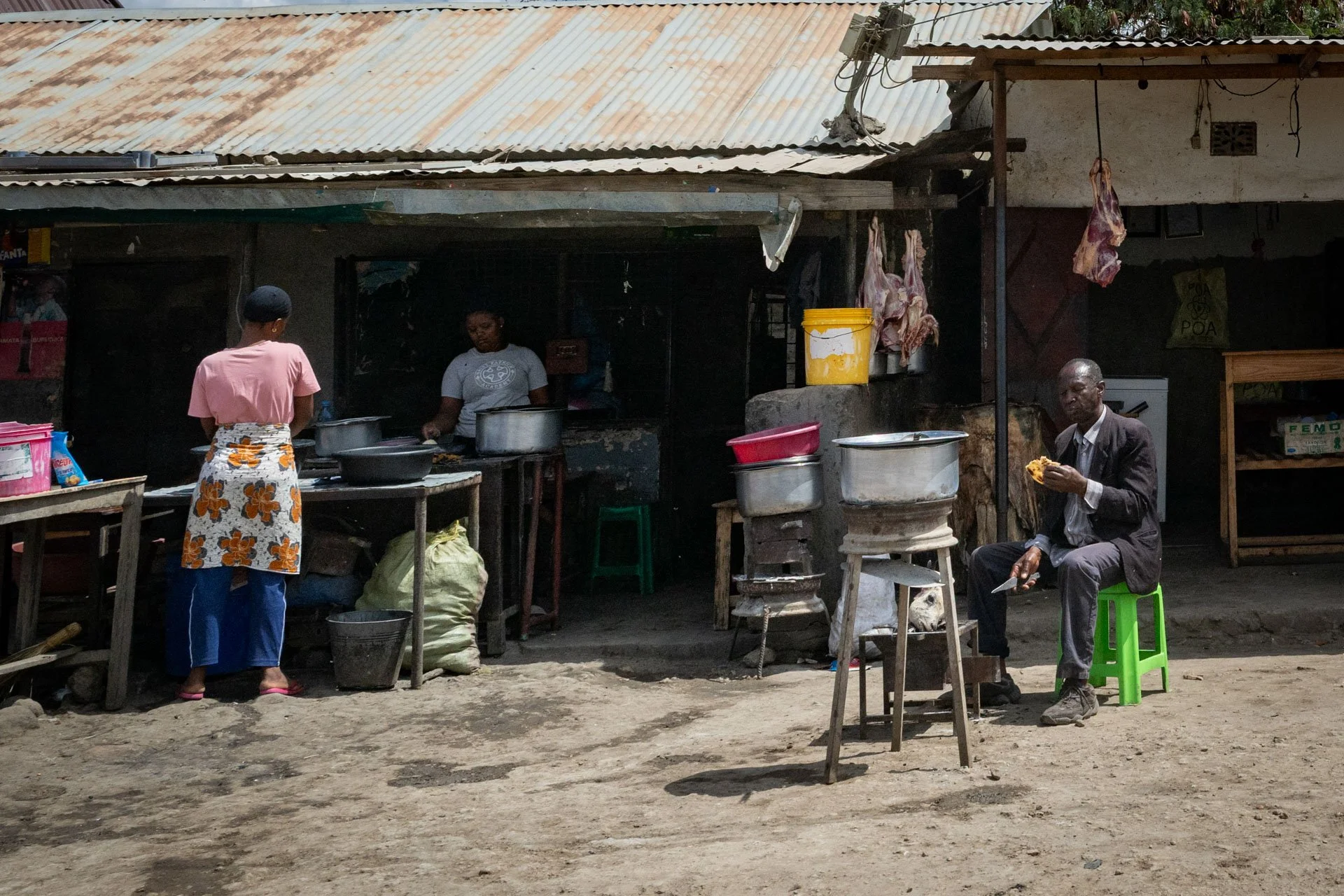 A street food stall with two women preparing food behind the counter and a man sitting on a green plastic stool eating. The stall has a rusted corrugated metal roof and hanging meat. The ground is dirt.