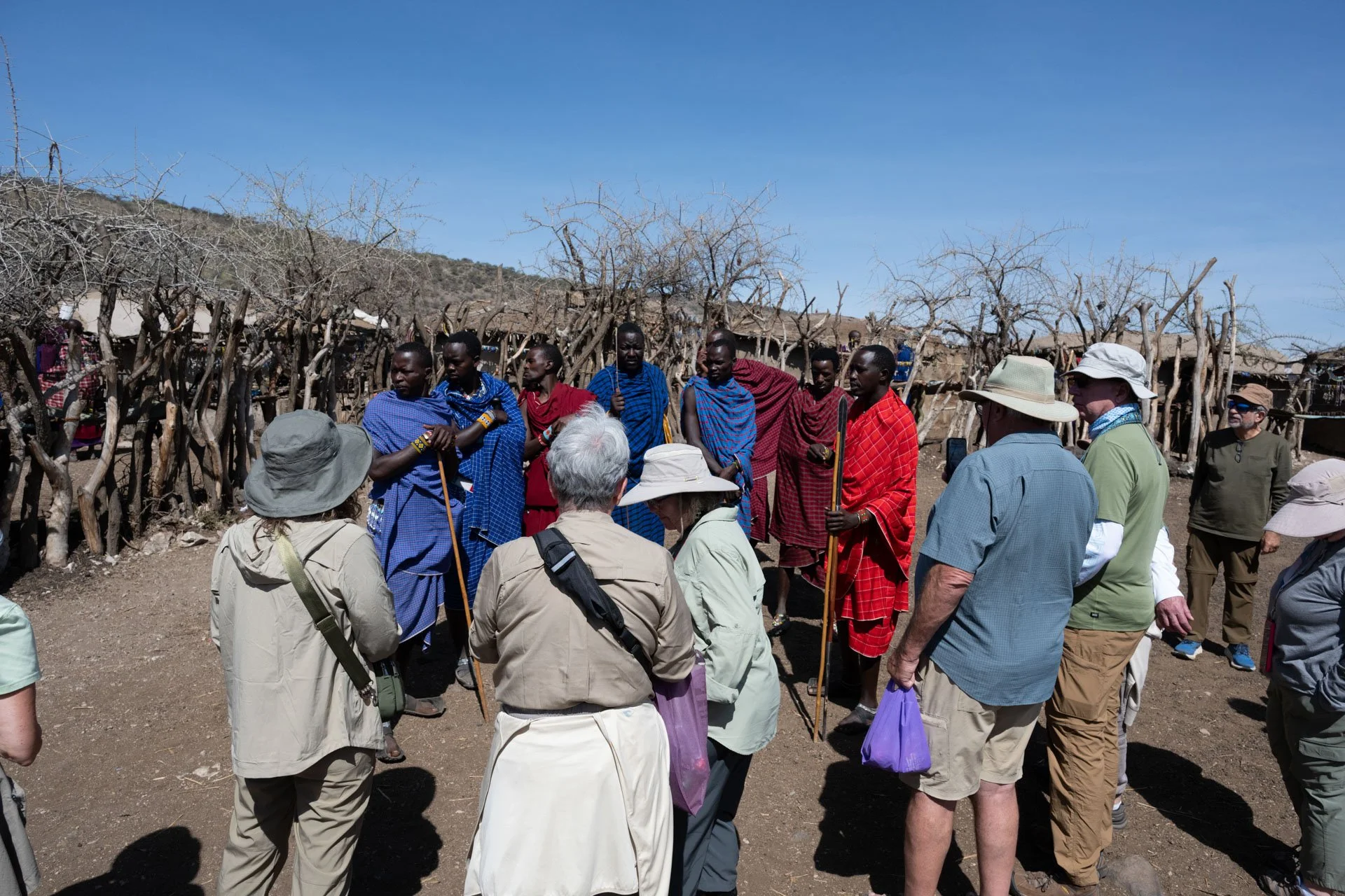 Group of tourists and Maasai people standing outdoors in a rural area with dry trees and simple structures, engaging in conversation.
